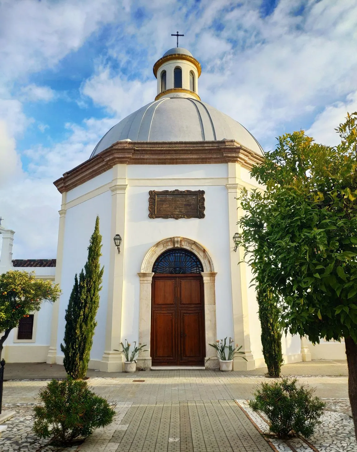 Capilla octogonal blanca con una cúpula gris rematada por una cruz, puerta de arco de madera y rodeada de cipreses y vegetación bajo un cielo azul