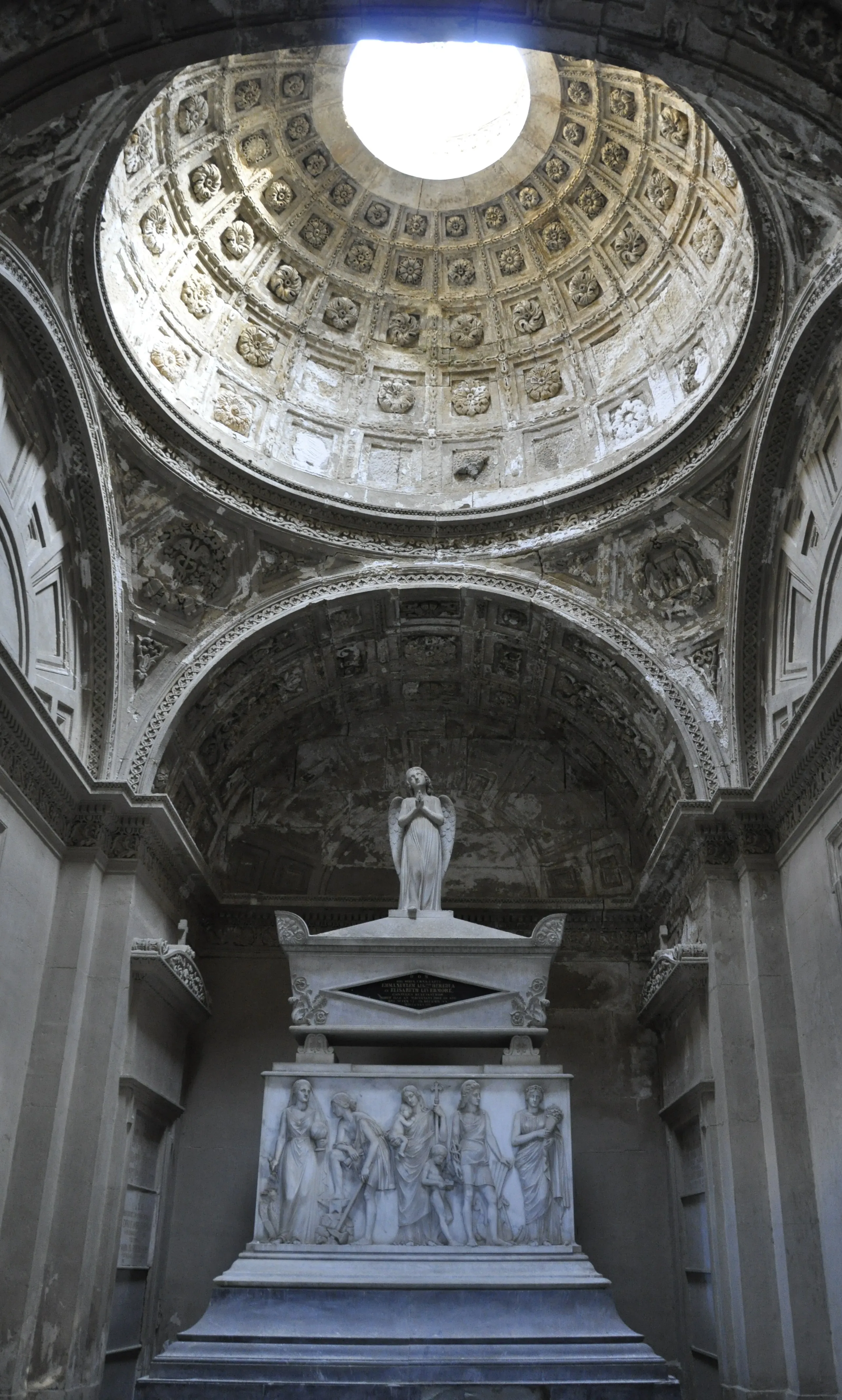 Vista interior de un mausoleo que muestra una cúpula artesonada con un óculo central sobre un sarcófago de mármol blanco rematado por una estatua de un ángel y relieves en su base.