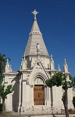 Witte stenen mausoleumkapel met een hoge, getrapte spits bekroond met een kruis, gotische boogdeur met ijzeren hek, tegen een helderblauwe lucht