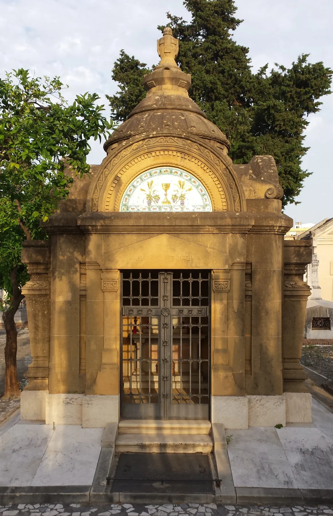 Mausoleo de piedra ornamentado con techo abovedado, panel de azulejos decorativos con floreros, verja de hierro con una cruz y una estatua sobre la cúpula, rodeado de árboles