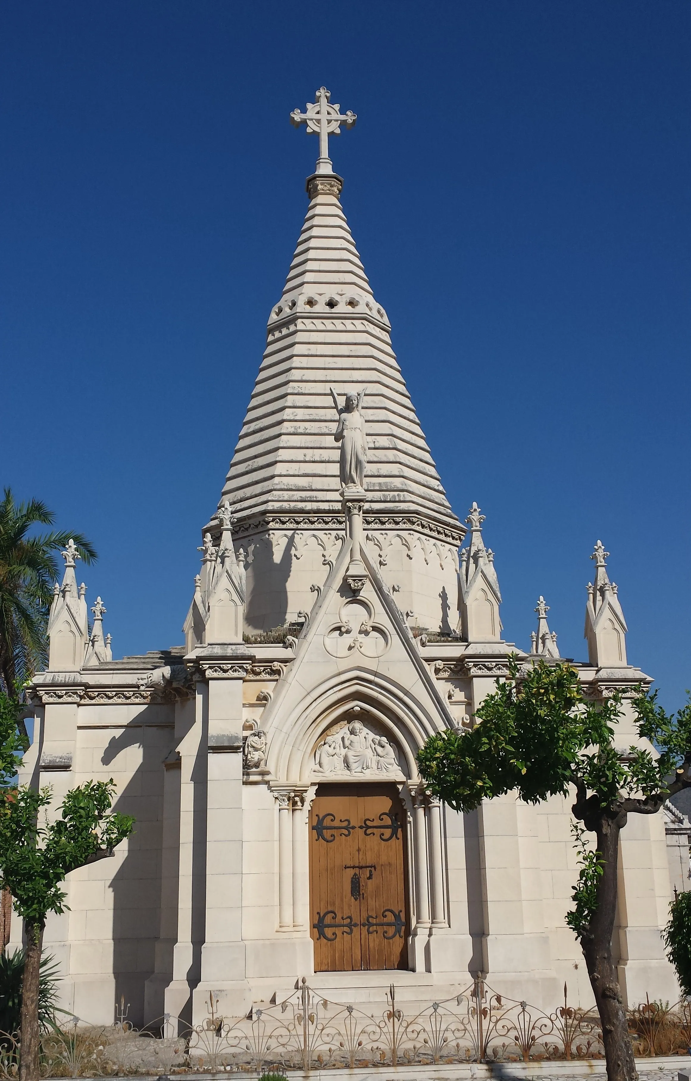 Mausoleo blanco de estilo gótico con una alta aguja cónica coronada por una cruz, una estatua de ángel y una puerta de madera arqueada en el Cementerio Histórico de San Miguel