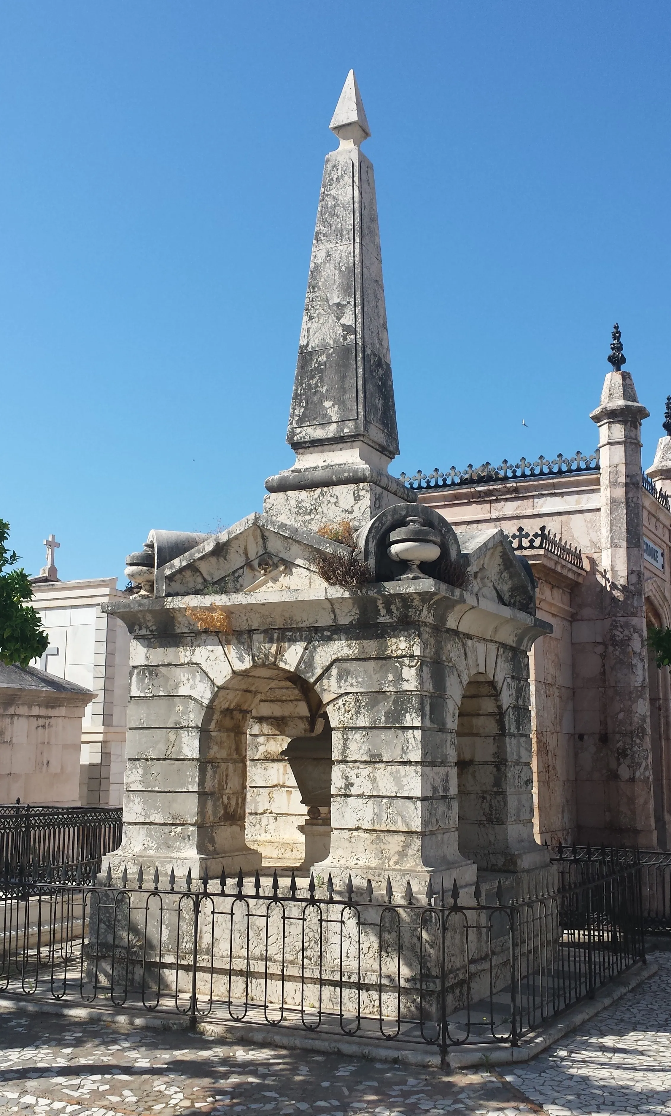 Mausoleo de piedra con un obelisco alto y nichos arqueados rodeado por una verja de hierro bajo un cielo azul despejado en el Cementerio Histórico de San Miguel.