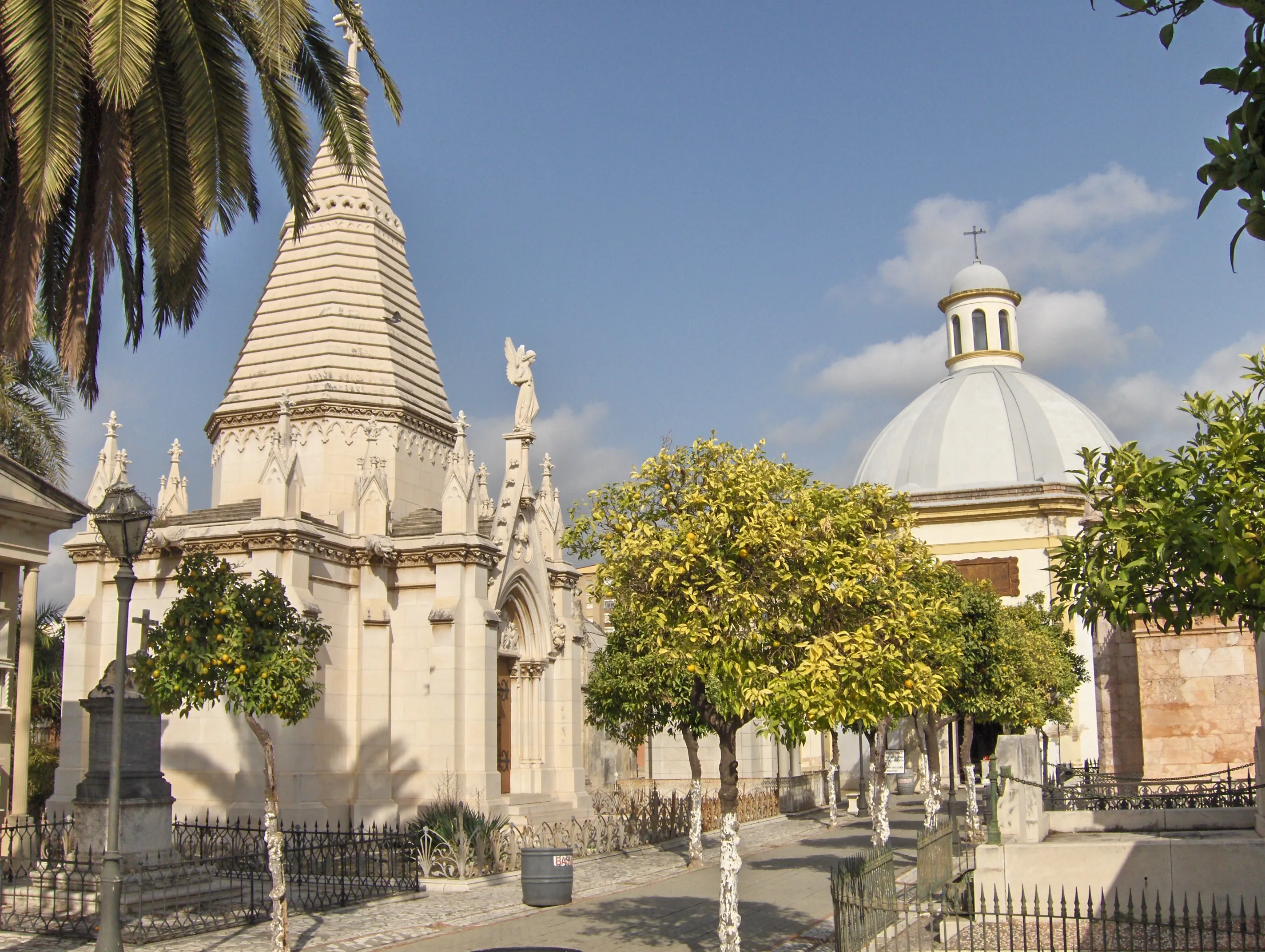Vista exterior del Cementerio Histórico de San Miguel que muestra un ornamentado mausoleo blanco con techo cónico y una estatua de un ángel junto a una capilla con cúpula bajo un cielo azul con palmeras