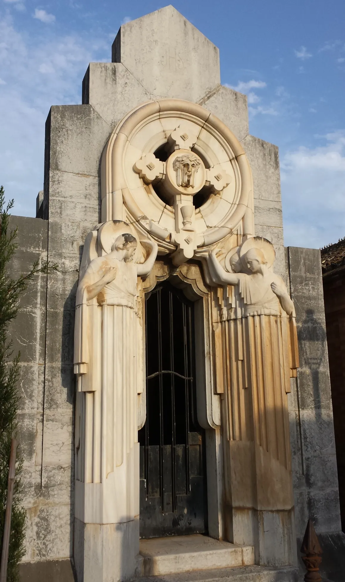 Fachada de mausoleo Art Déco con dos ángeles esculpidos flanqueando una puerta de hierro negro, rematada por un medallón de piedra circular con un motivo de calavera, sobre un cielo azul