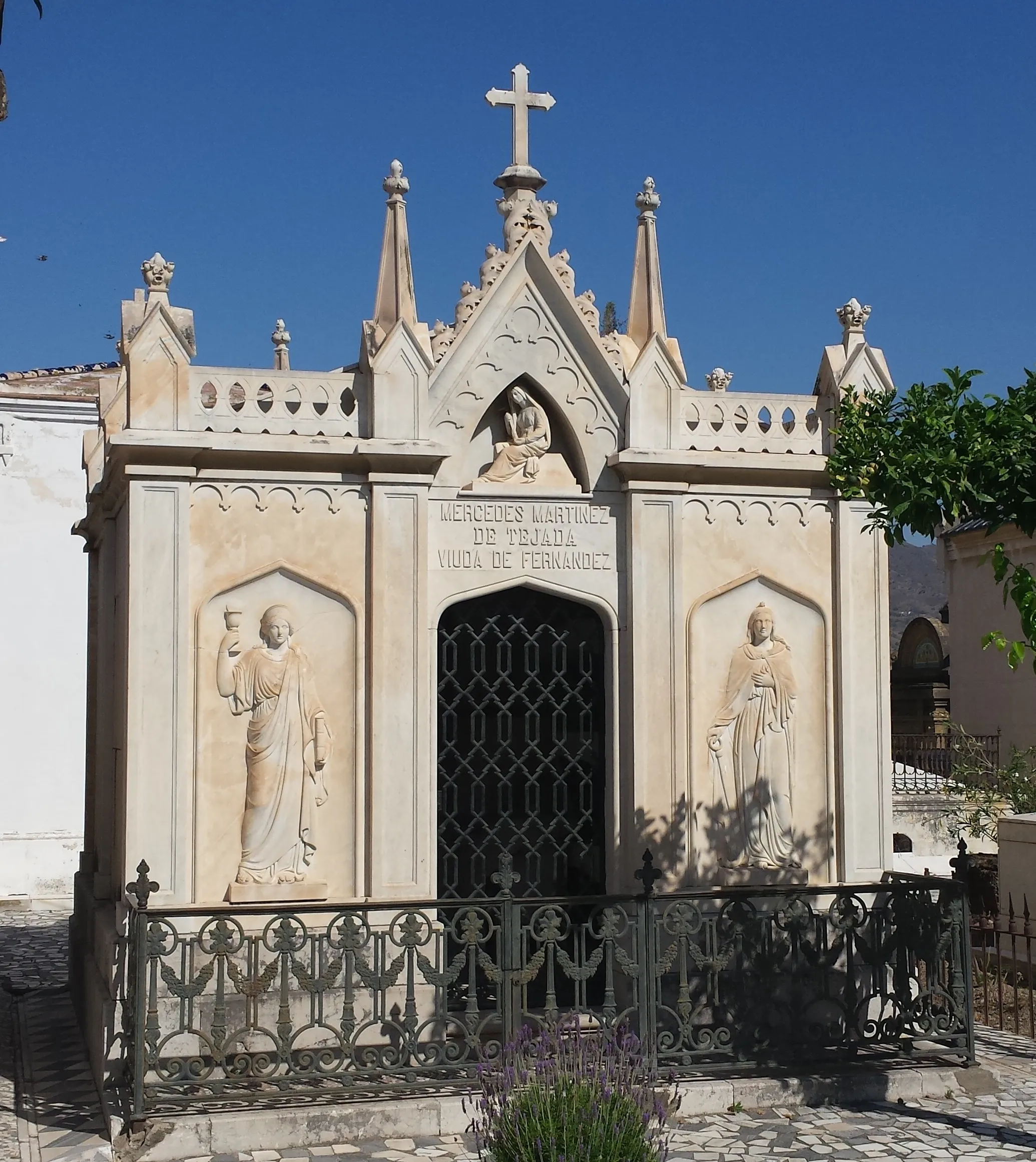 Mausoleo neogótico con una cruz central sobre agujas puntiagudas, flanqueado por dos esculturas en relieve de figuras con túnicas, una entrada con puerta de hierro arqueada y una placa de dedicación con inscripciones, rodeado por una verja de hierro decorativa bajo un cielo azul despejado.