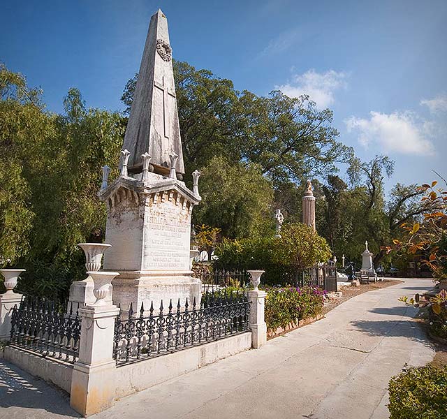 Obelisco de piedra como monumento funerario con verja de hierro negro a lo largo de un sendero en el Cementerio Inglés de Málaga, rodeado de árboles y otros monumentos bajo un cielo azul.