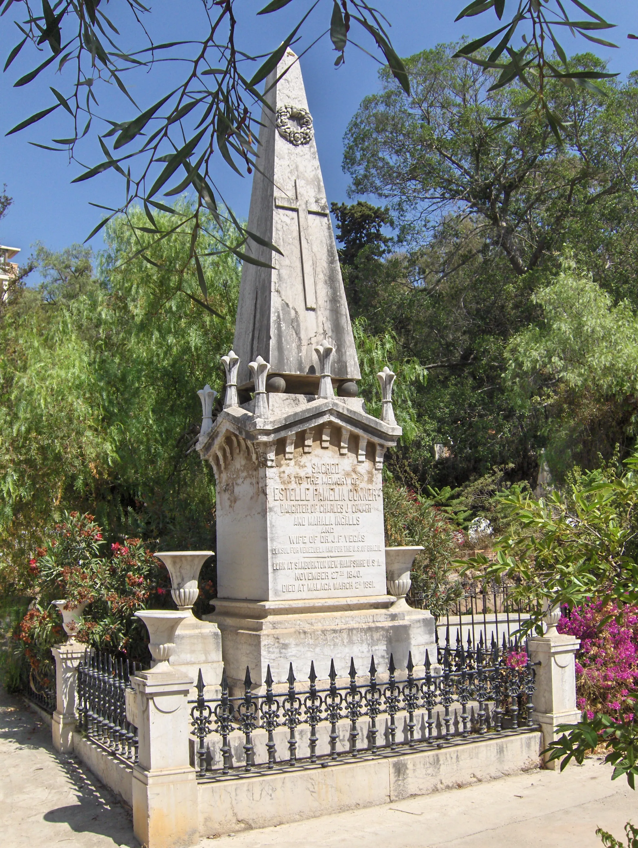 Alto monumento funerario de obelisco de piedra blanca con una cruz y una placa inscrita, rodeado por una reja de hierro negro, entre árboles verdes y flores de buganvilla rosa bajo un cielo azul