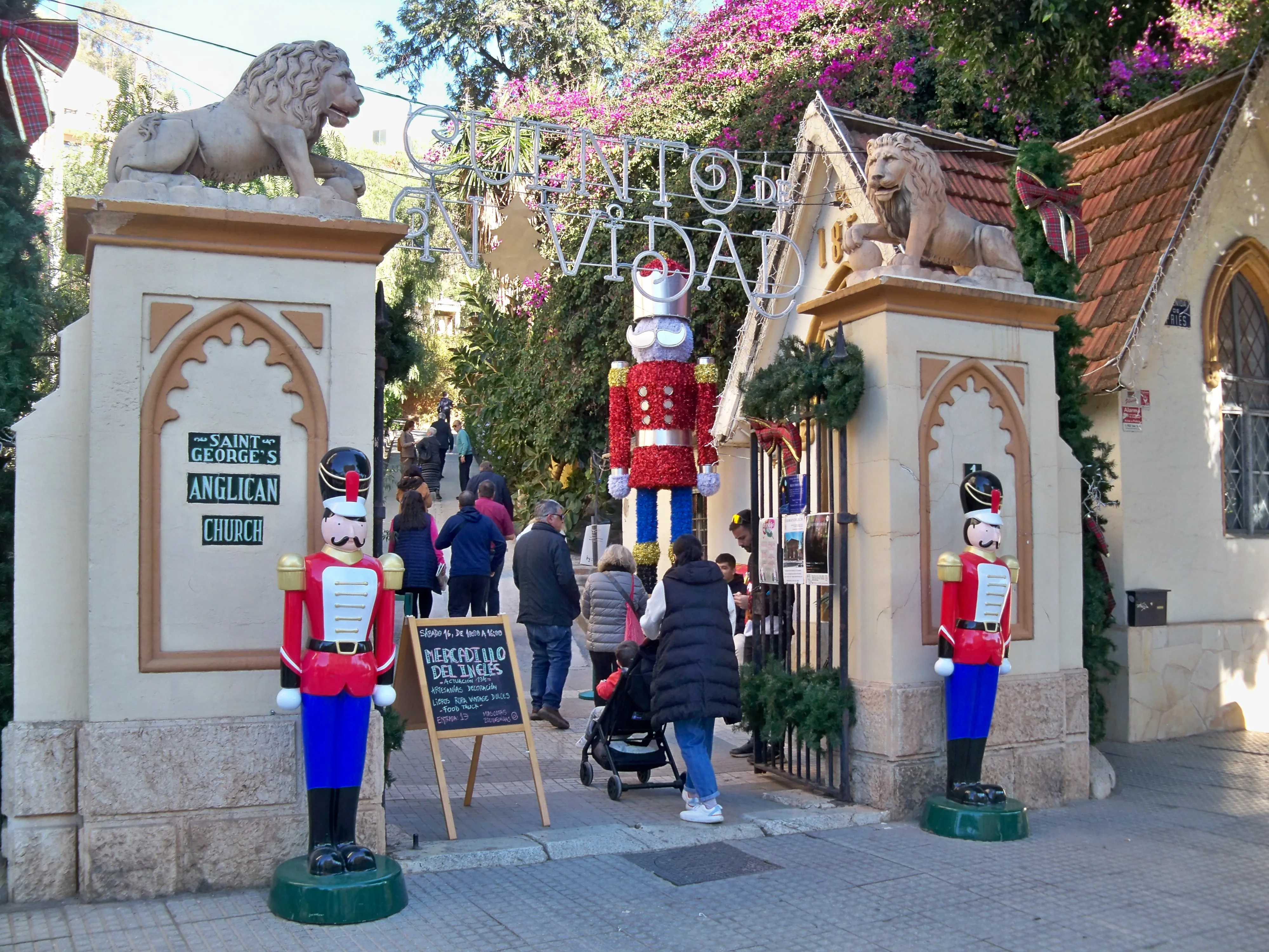 Puerta de entrada del Cementerio Inglés de Málaga con estatuas de leones en los pilares, grandes figuras de soldados cascanueces y visitantes caminando a través del arco