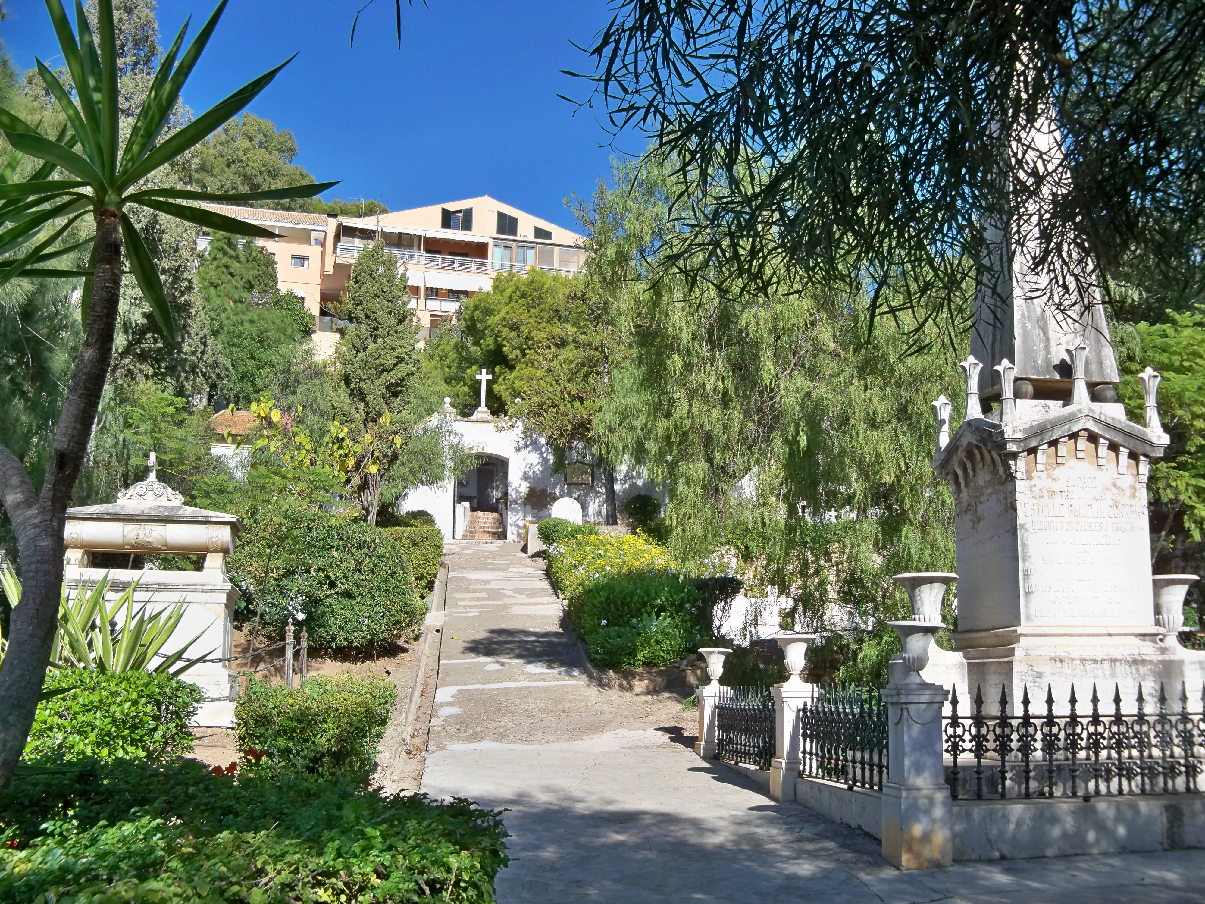 Un sendero soleado bordeado de monumentos funerarios de piedra blanca y árboles verdes conduce a una puerta de entrada arqueada en el Cementerio Inglés de Málaga, con edificios residenciales visibles en la ladera de arriba.