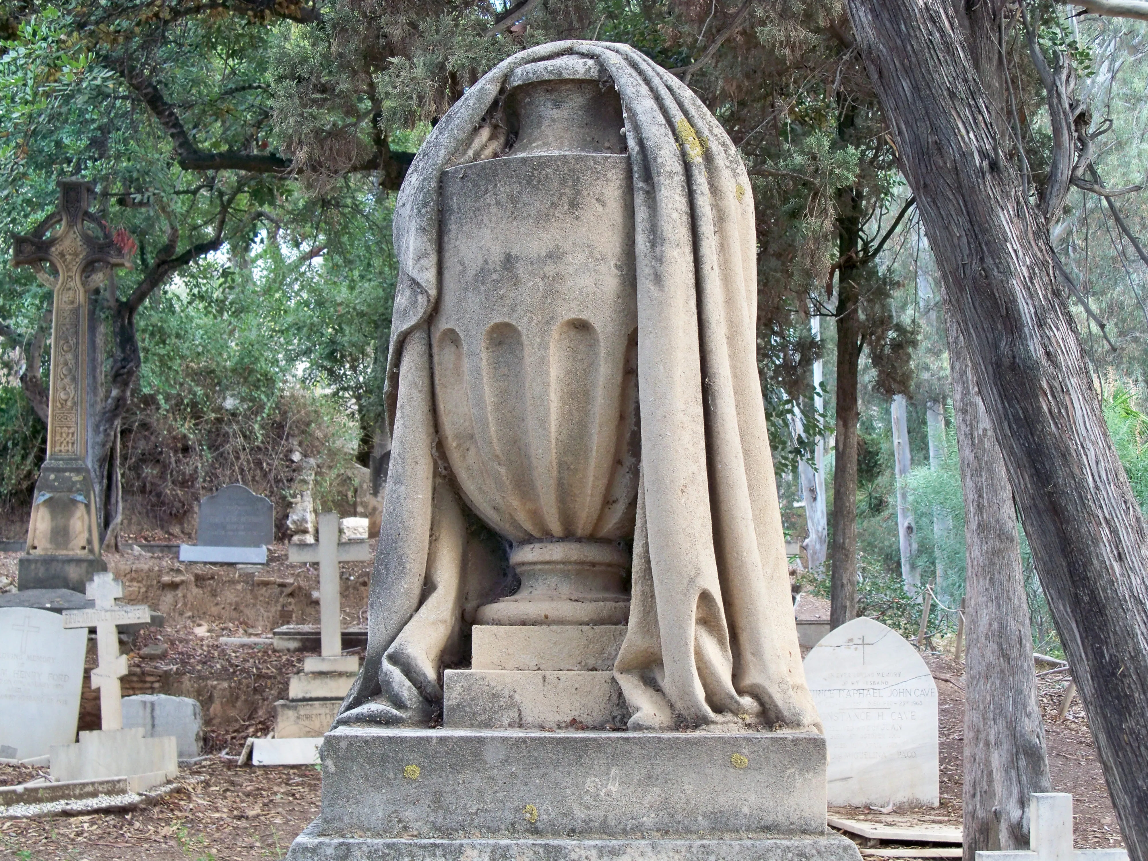 Urna funeraria de piedra cubierta con un sudario tallado sobre un pedestal, rodeada de lápidas, un monumento en forma de cruz y árboles en un cementerio.