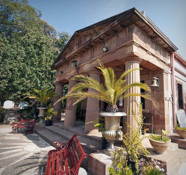 Edificio clásico de piedra con columnas y un frontón triangular, flanqueado por palmeras en grandes maceteros de urna, con bancos de hierro rojo en un patio bañado por el sol