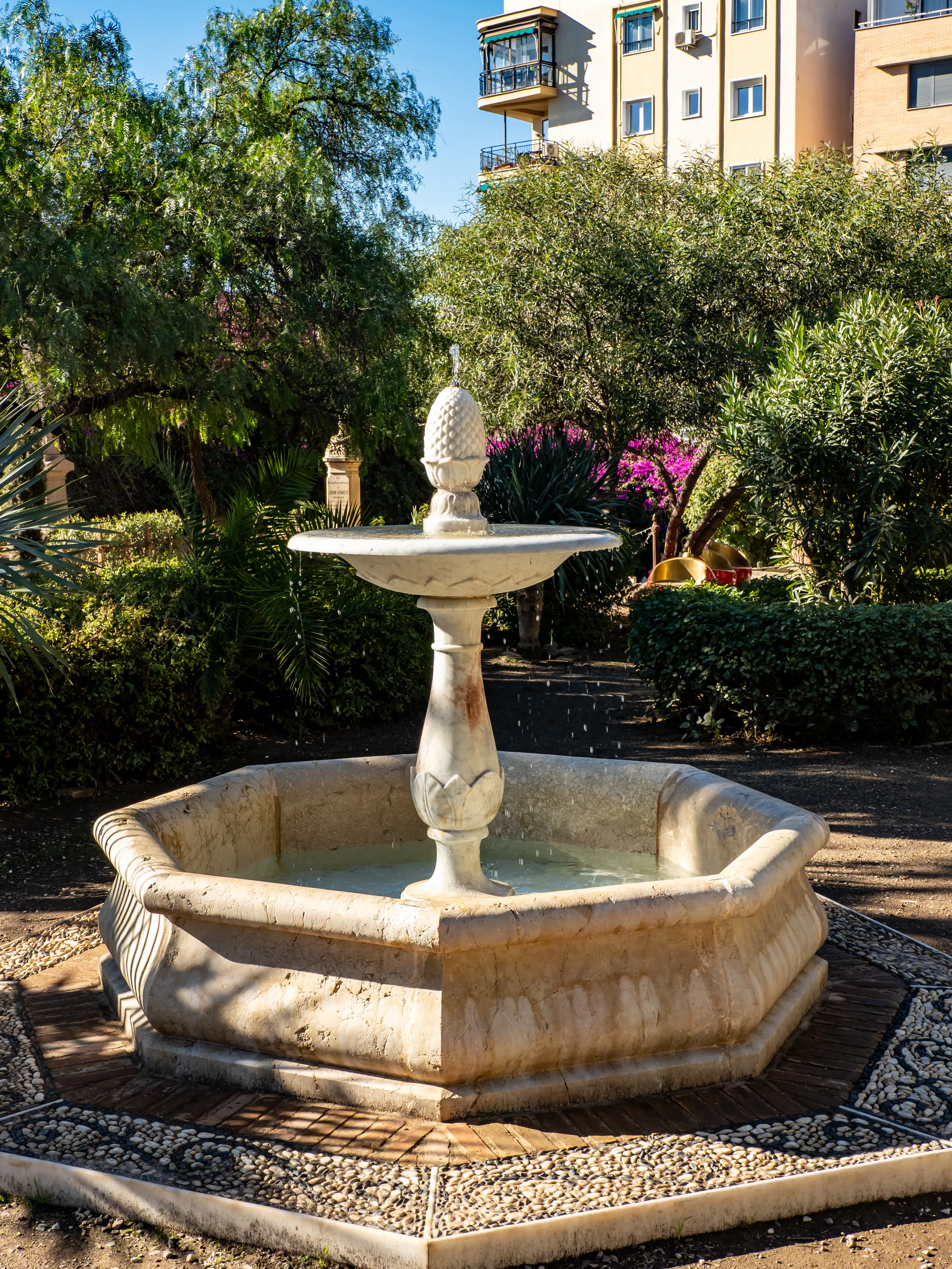 Fuente de piedra con remate en forma de piña en el jardín del Cementerio Inglés de Málaga, rodeada de árboles verdes y arbustos de flores moradas con un edificio visible al fondo.