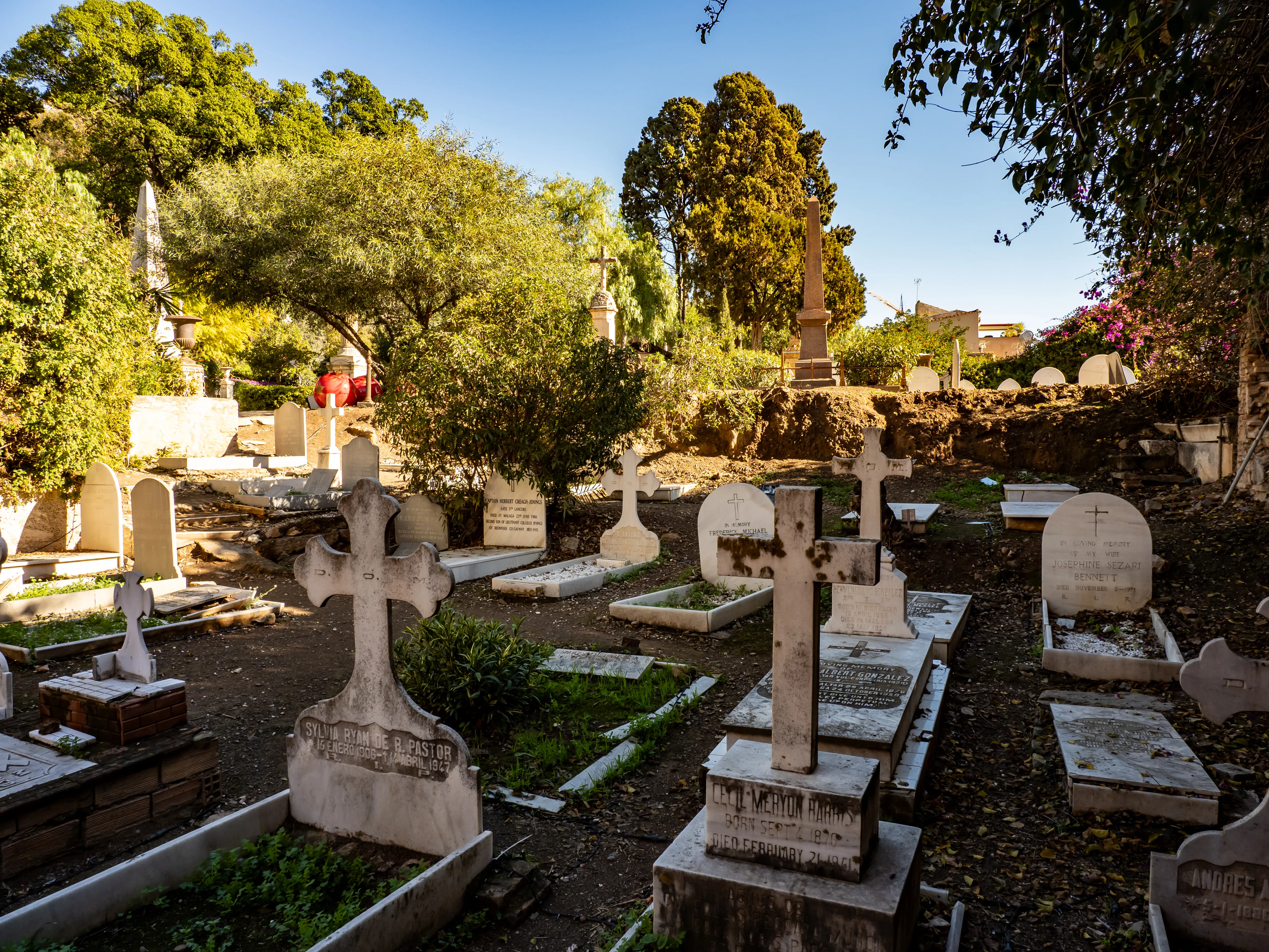 Vista exterior del Cementerio Inglés de Málaga mostrando numerosas lápidas de piedra blanca, cruces y monumentos entre árboles verdes bajo un cielo azul despejado.