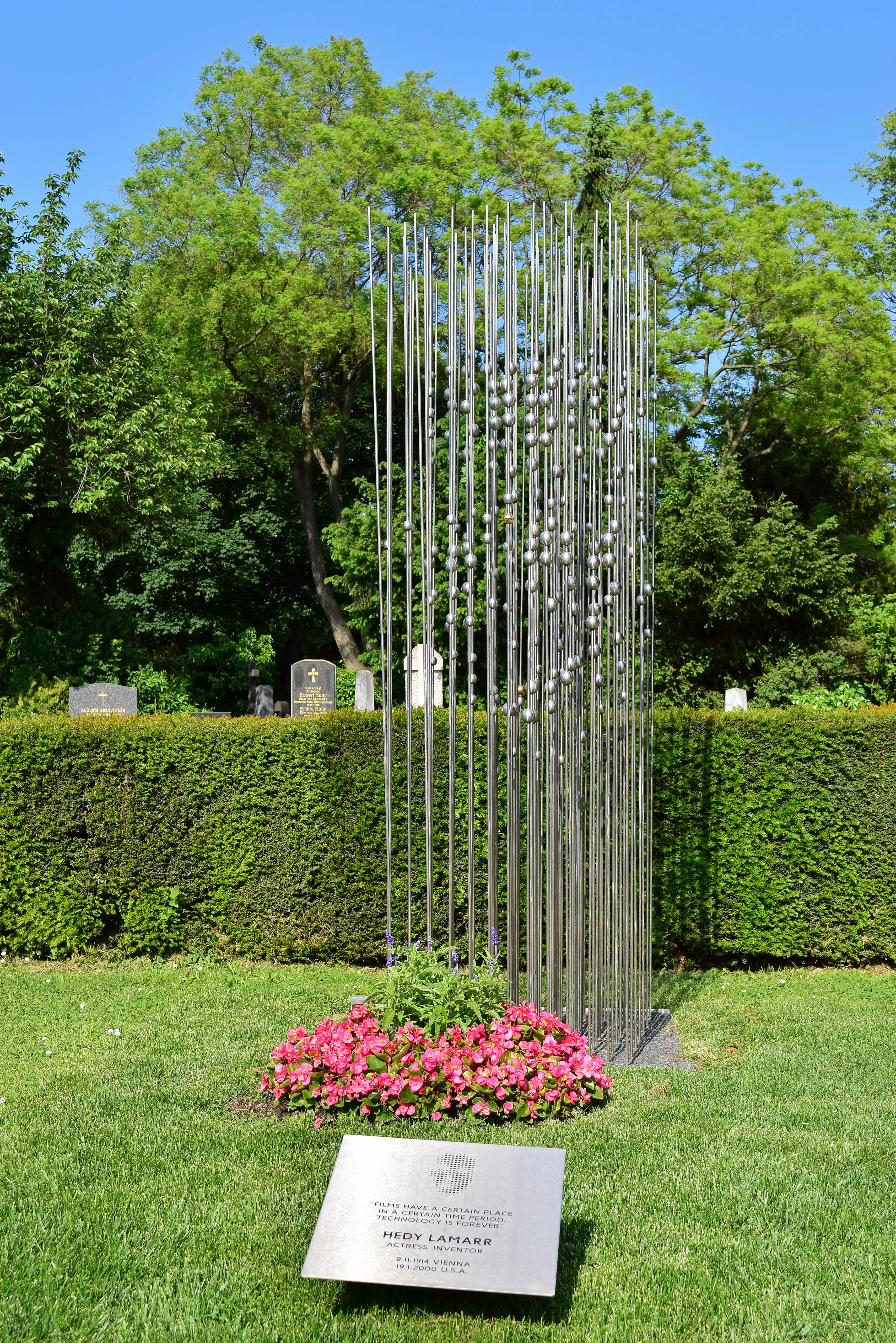 Memorial to Hedy Lamarr with a modern sculpture of vertical metal rods and spherical elements, surrounded by pink flowers and a plaque, in a green cemetery setting.