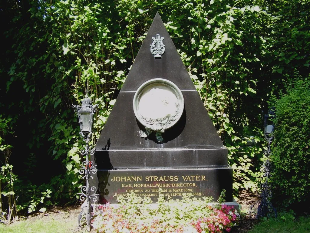 Monumento triangular negro con un medallón circular y el texto 'JOHANN STRAUSS VATER' rodeado de vegetación y flores