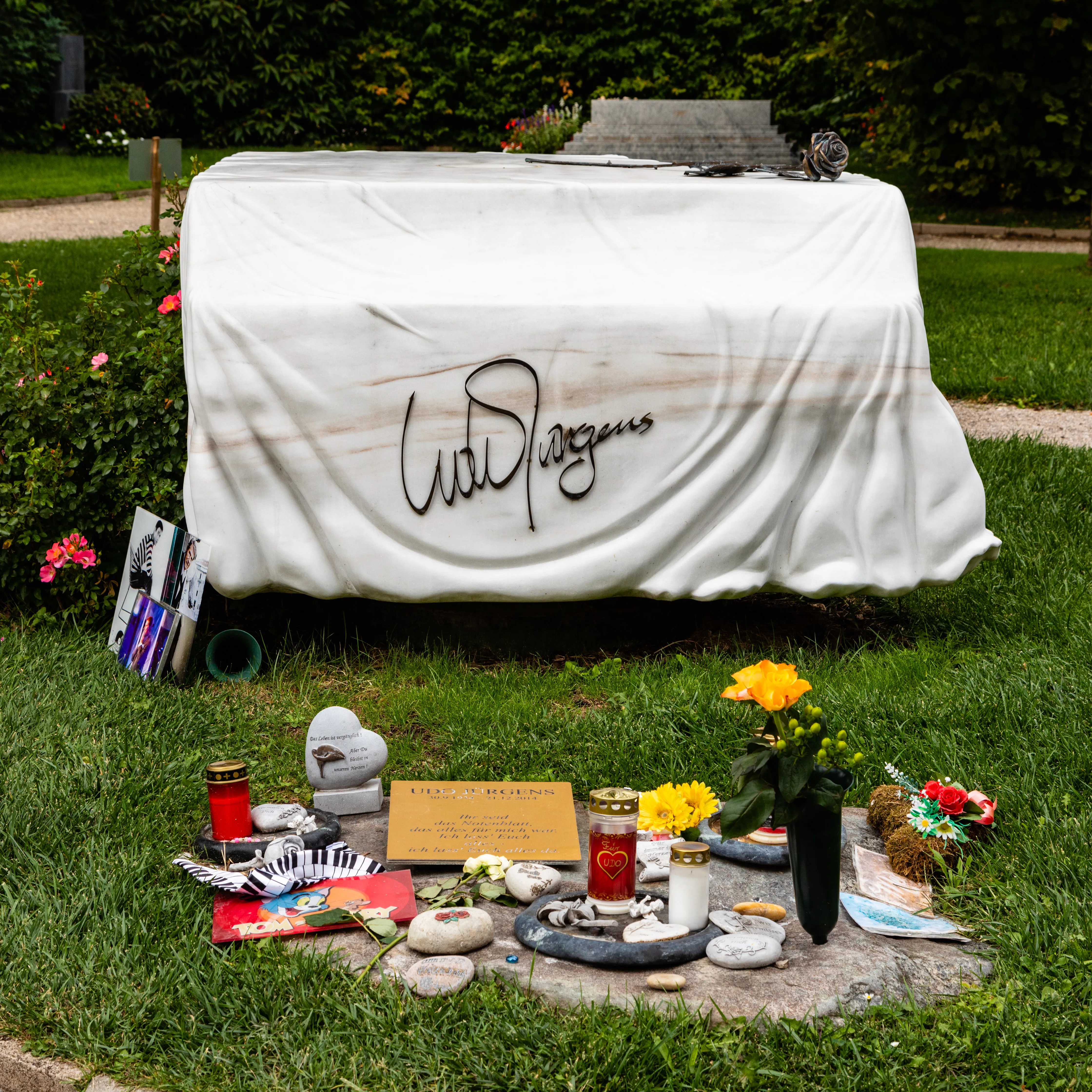 Una tumba cubierta con una tela blanca que presenta la firma 'Udo Jürgens', rodeada de flores, velas y objetos personales en una zona de césped.