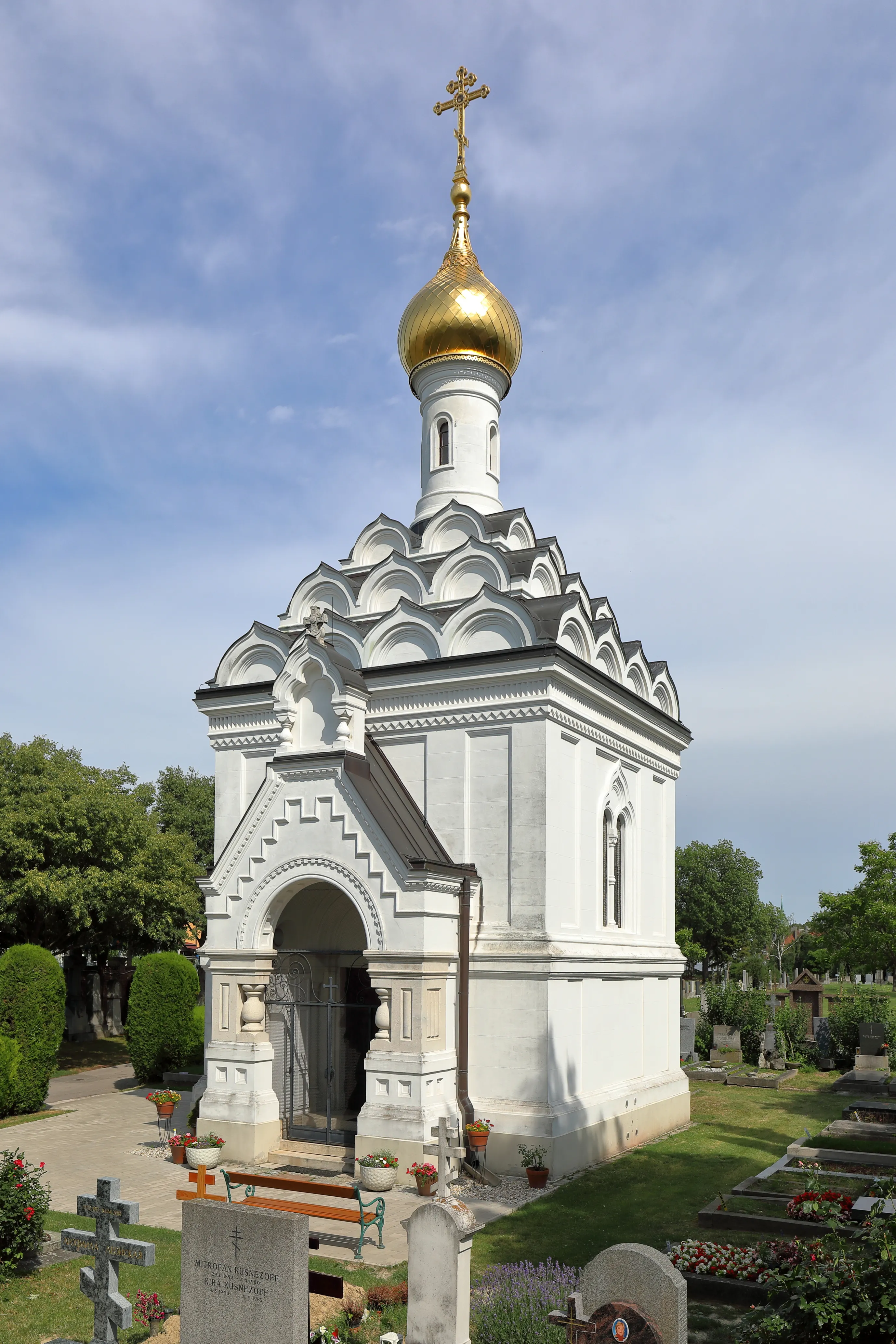 A white stone chapel with a golden onion dome and cross, surrounded by greenery and tombstones in a cemetery.
