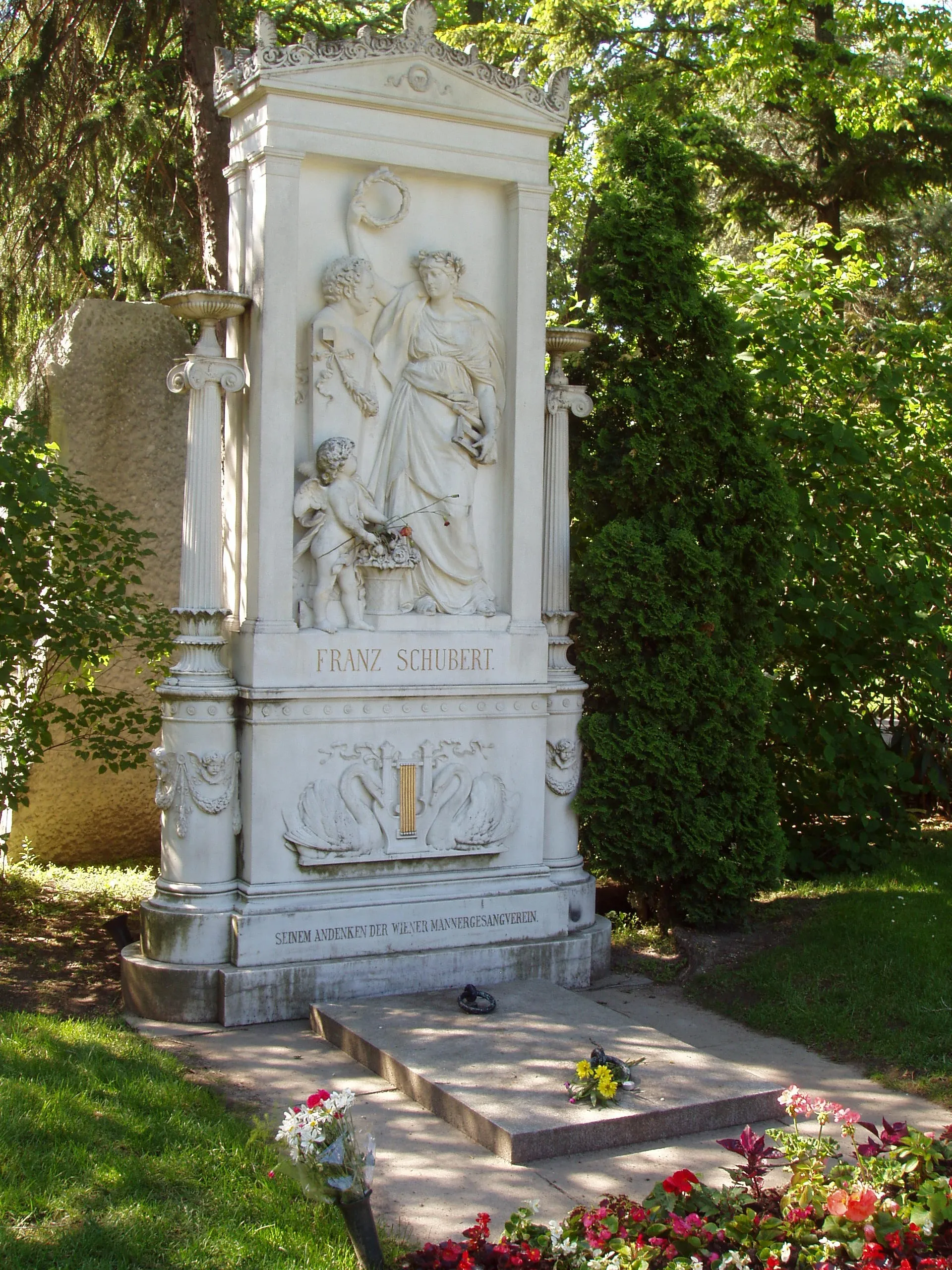 White marble monument with sculpted figures and the name Franz Schubert, surrounded by greenery and flowers