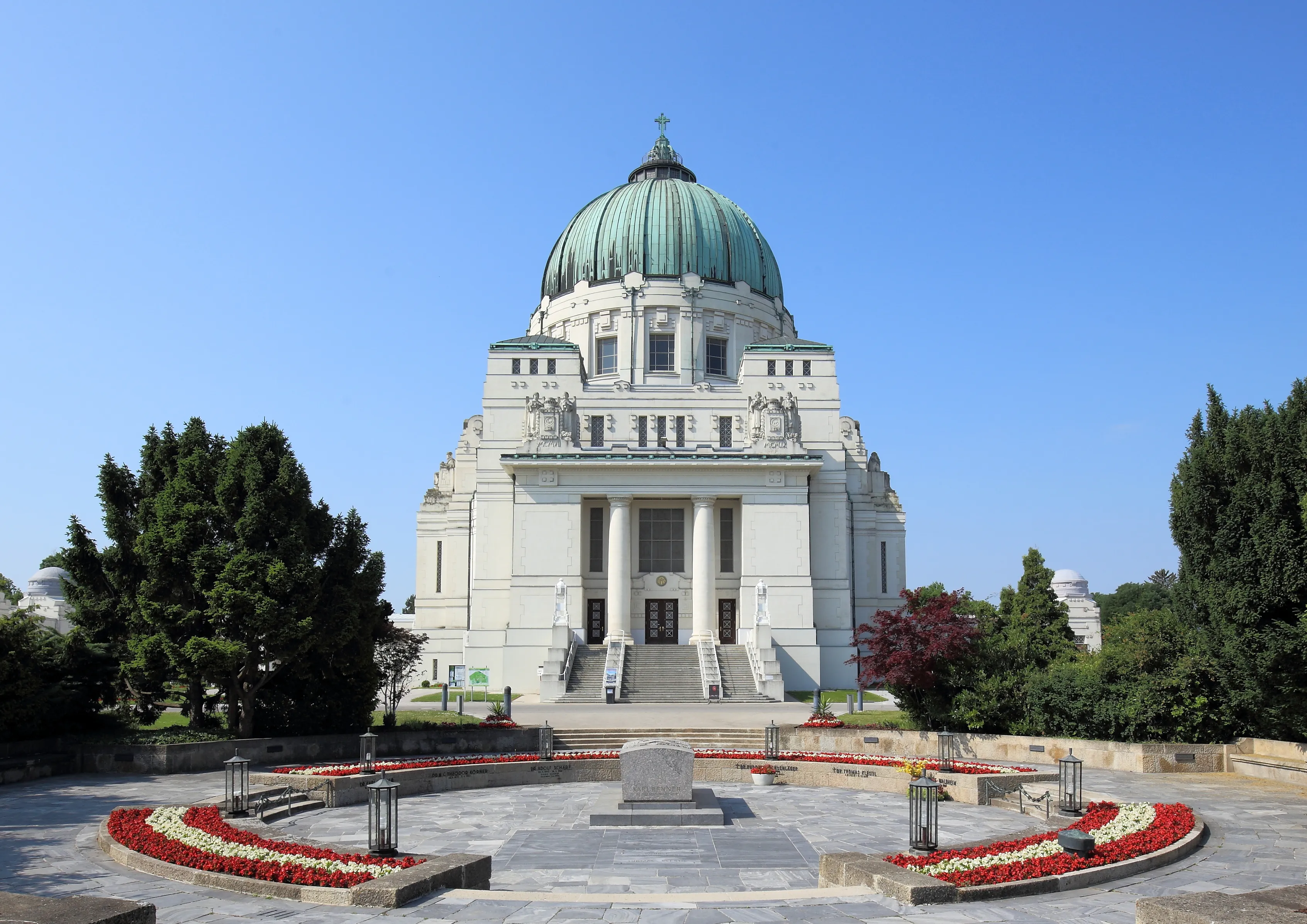 A large white building with a green dome and classical columns, surrounded by trees and a circular flowerbed with red and white flowers under a clear blue sky