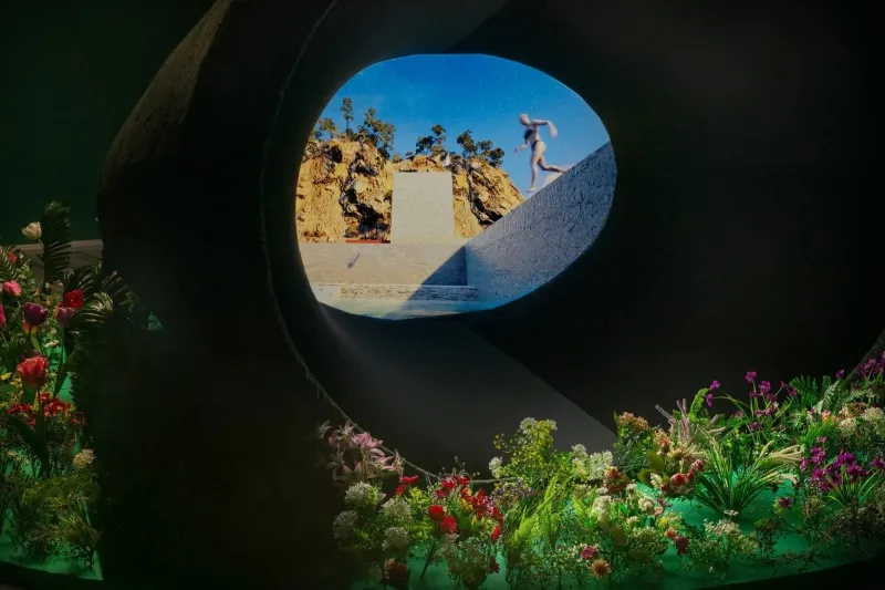 Abertura circular en una estructura oscura que revela la imagen proyectada de una figura en un paisaje rocoso, rodeada de flores y plantas.