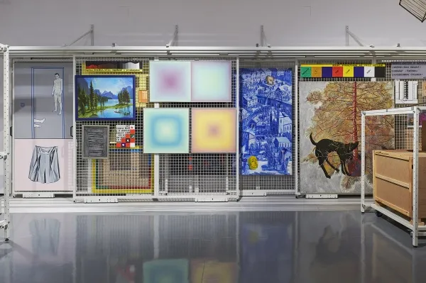 Una galería con diversas obras de arte expuestas detrás de una reja metálica, que incluyen pinturas, pantallas digitales y un gran mural con la ilustración de un perro.