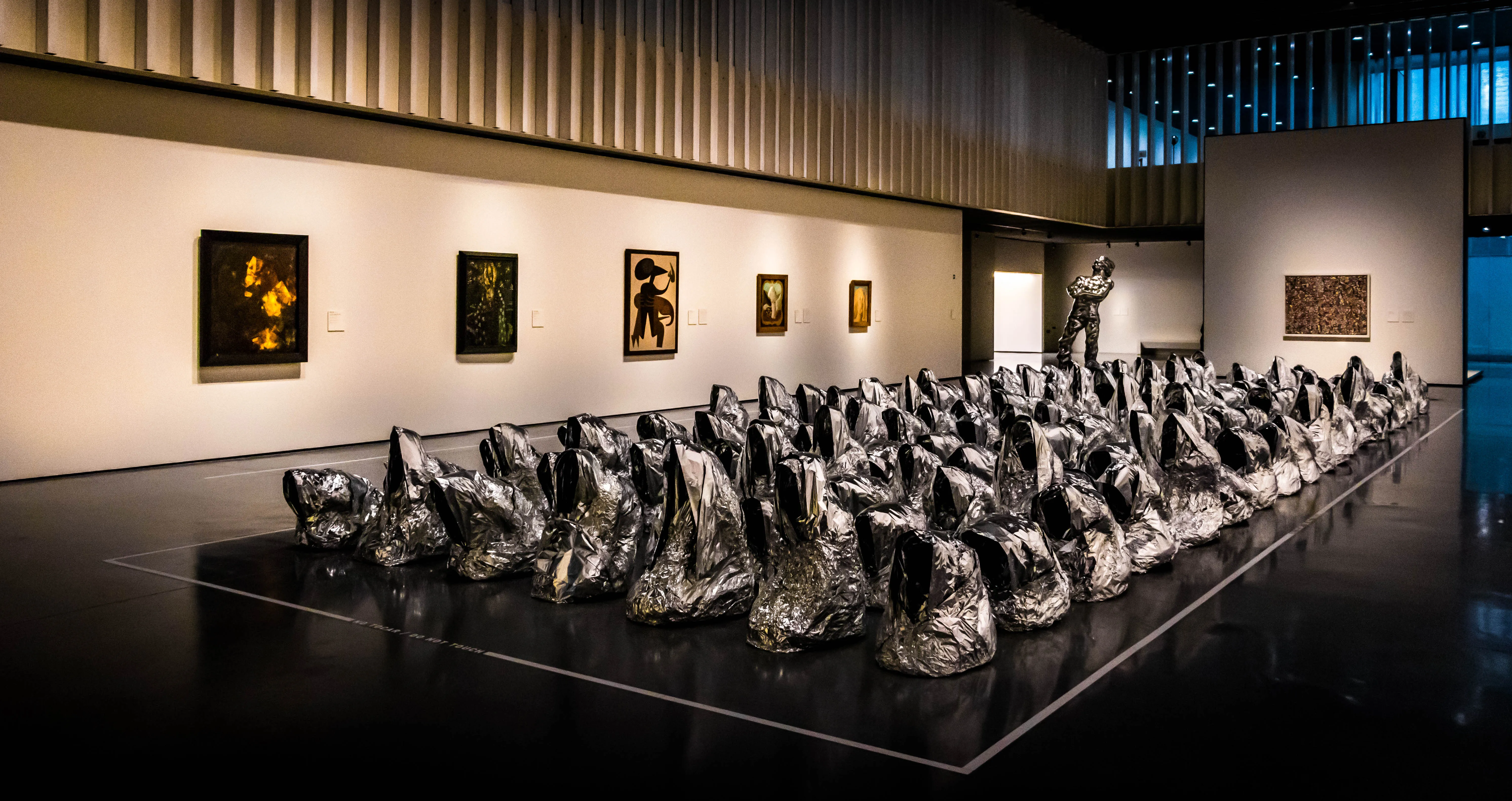 Interior de una galería de museo con una gran instalación de formas escultóricas metálicas esparcidas por un suelo oscuro pulido, con pinturas enmarcadas colgadas en paredes blancas bajo un techo de lamas