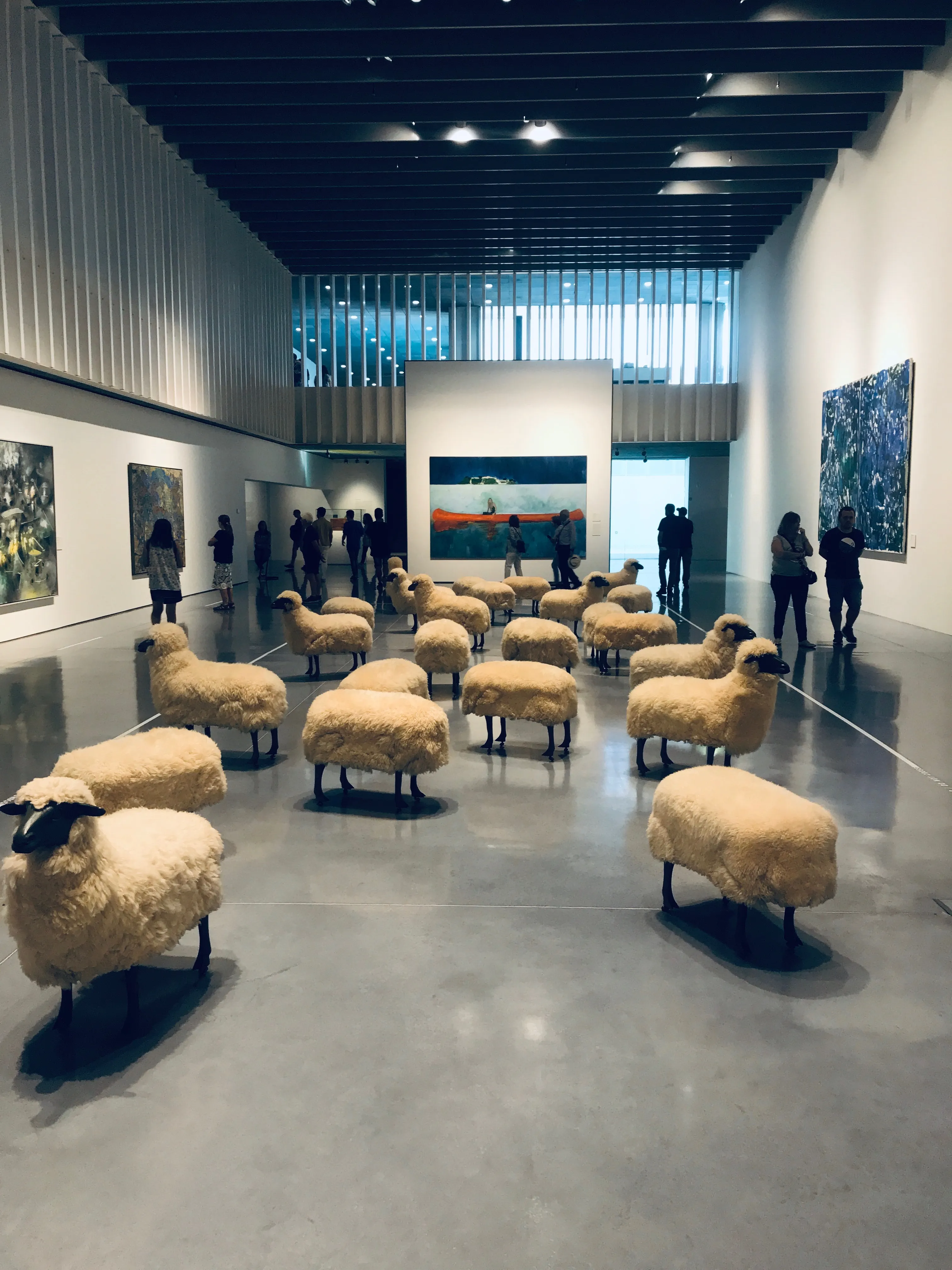 Interior de una galería de arte que presenta una instalación de esculturas realistas de ovejas dispuestas en el suelo, con visitantes observando cuadros en las paredes.