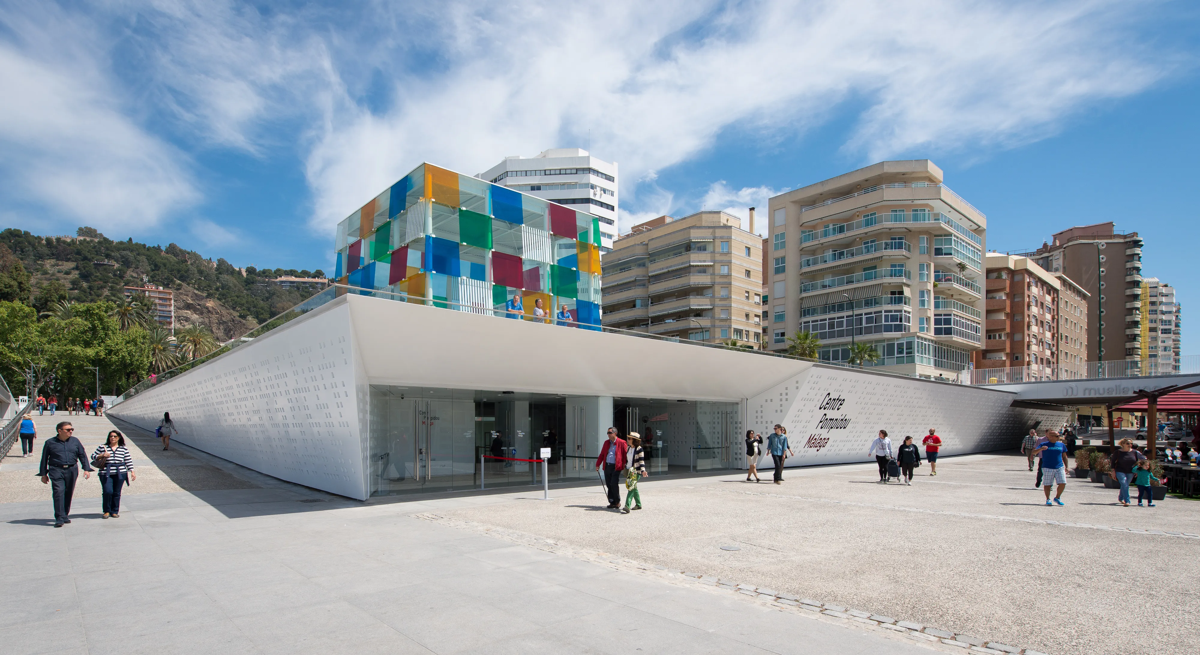 Vista exterior del Centre Pompidou Málaga que muestra una estructura cúbica multicolor sobre un edificio blanco de formas angulares, con personas caminando en la plaza y edificios urbanos circundantes al fondo.