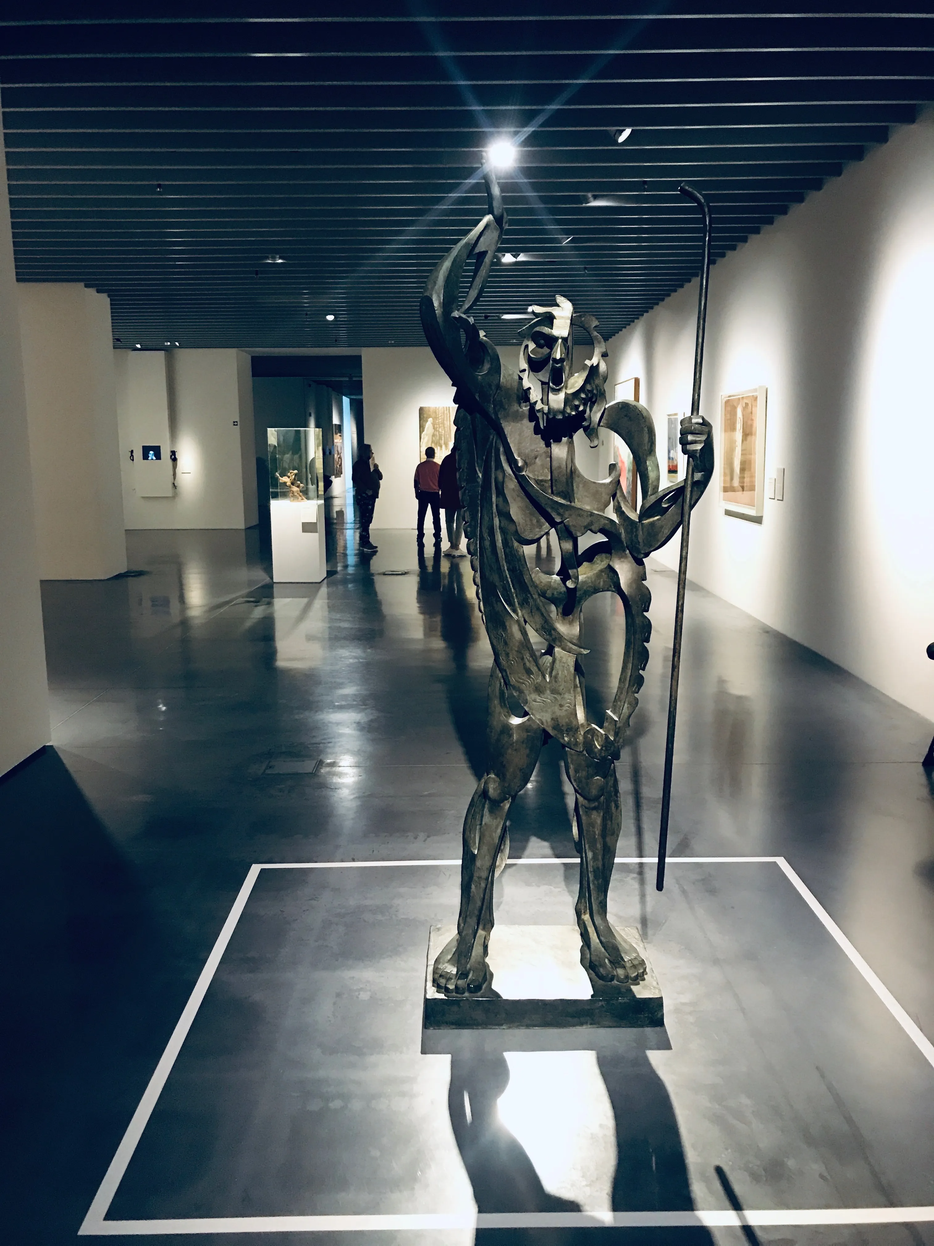 Escultura de bronce de una figura tipo centauro con un brazo levantado sosteniendo un bastón, de pie sobre un pedestal en una sala de museo con visitantes al fondo