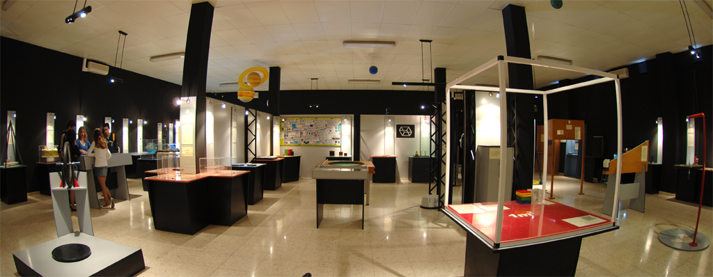 Vista panorámica interior de una sala de exposiciones de un centro de ciencias con mesas interactivas, vitrinas de cristal, paredes oscuras, suelos claros, focos en el techo y visitantes explorando las exposiciones