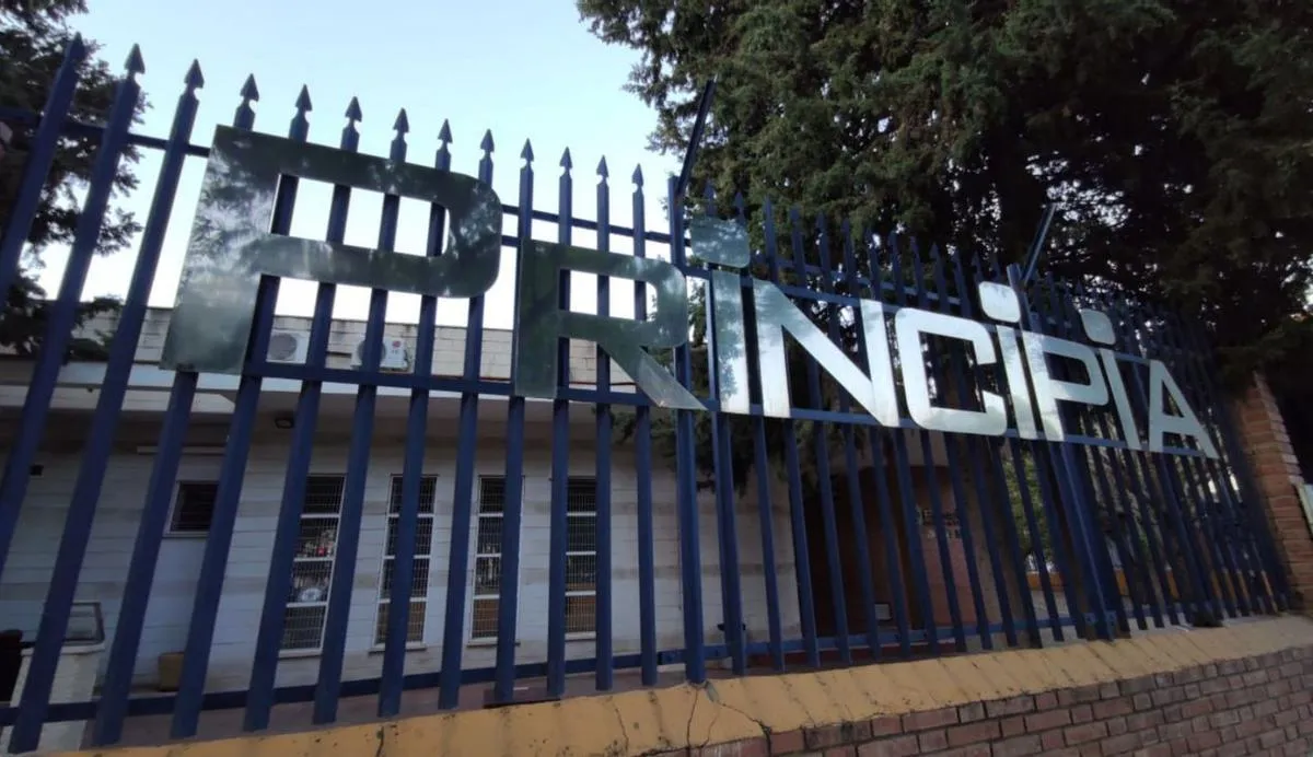 Valla metálica azul con grandes letras de metal que forman la palabra PRINCIPIA montadas sobre ella, con un edificio y árboles visibles detrás.