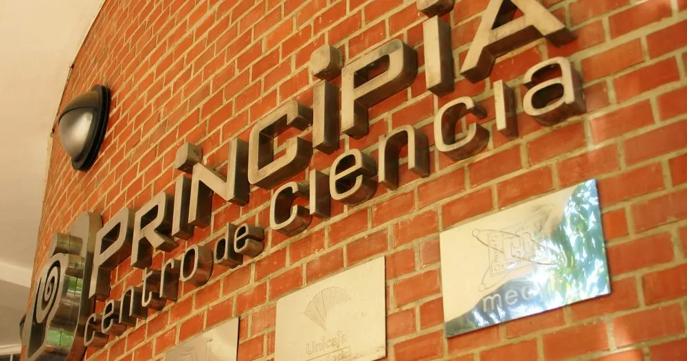 Vista en ángulo de una pared de ladrillo rojo que presenta grandes letras metálicas en 3D con el texto 'PRINCIPIA centro de ciencia' y placas informativas montadas debajo.