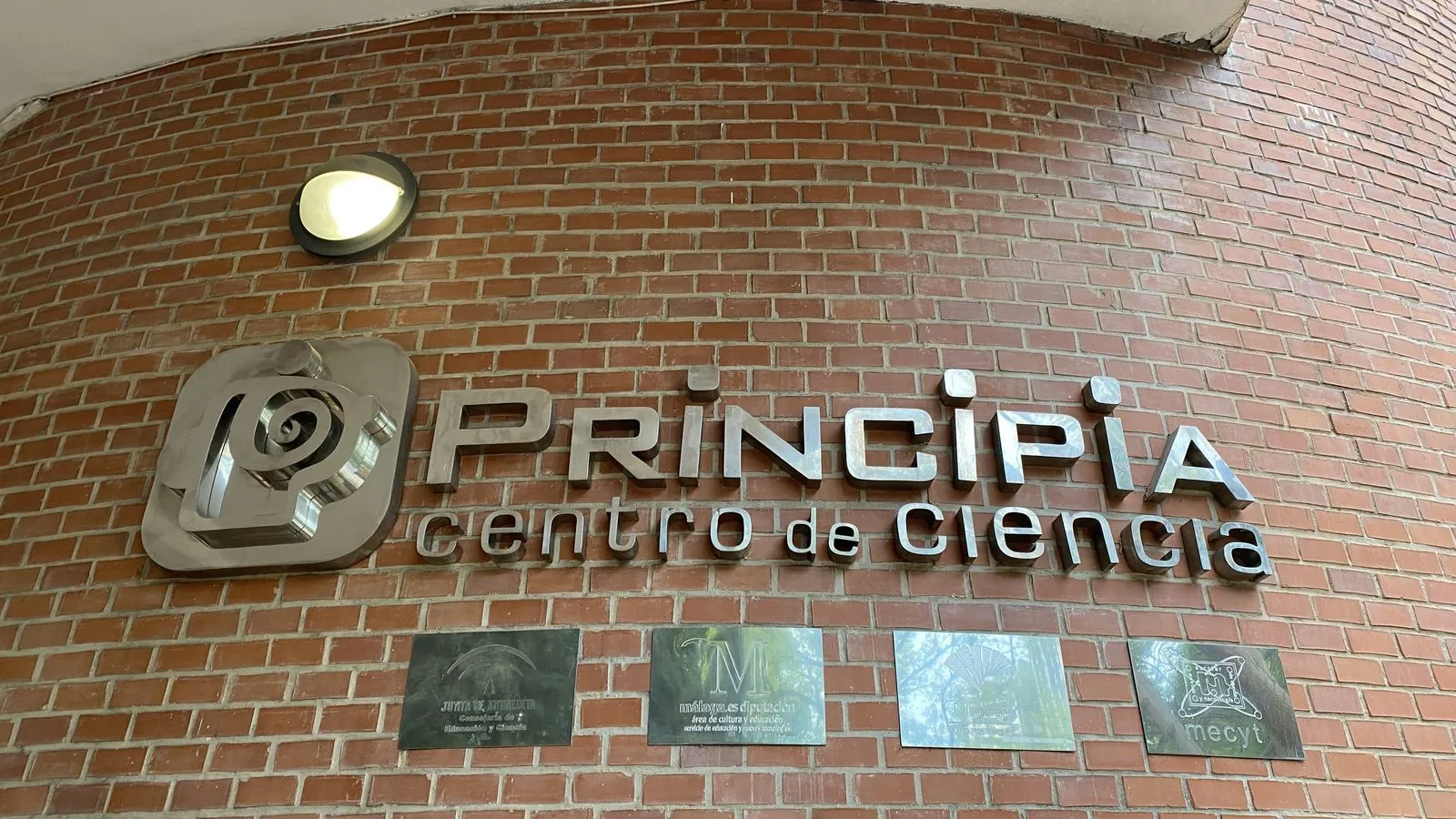 Fachada de ladrillo con un gran letrero metálico y logotipo de 'PRINCIPIA centro de ciencia', con cuatro placas conmemorativas montadas debajo