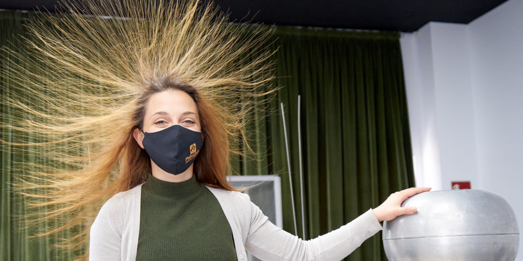 Mujer con el pelo erizado por la electricidad estática, tocando una gran esfera plateada de un generador de Van de Graaff mientras lleva una mascarilla oscura, con un telón de fondo de cortina verde