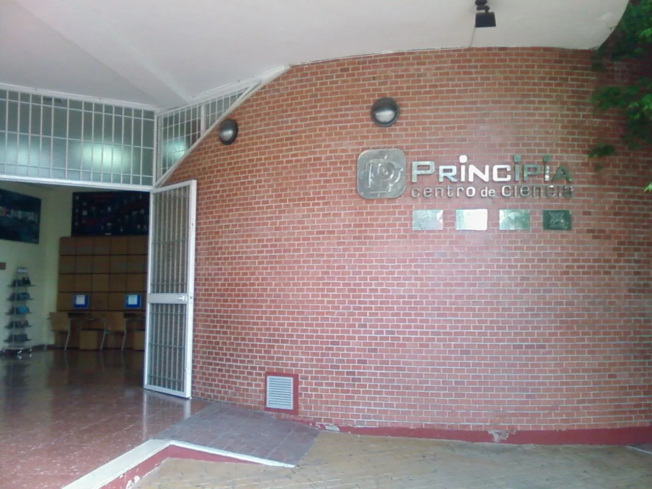 Vestíbulo de entrada del Principia Centro de Ciencia con una pared de ladrillo curva que muestra el nombre del lugar y el logotipo, y una puerta metálica abierta que conduce a una sala interior con estaciones de ordenador.