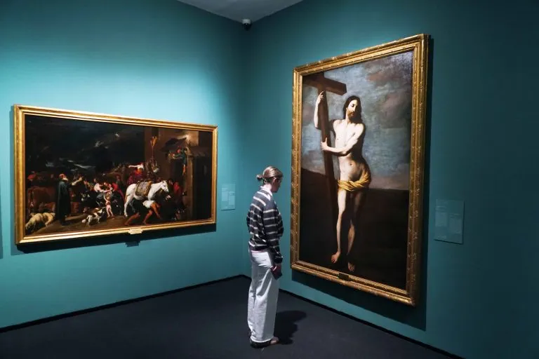 Dos grandes cuadros con marcos dorados sobre paredes de galería de color verde azulado, con un visitante con una camiseta de rayas de pie entre ellos observando la obra de arte