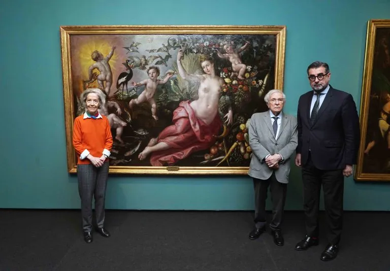 Tres personas posan frente a un gran cuadro clásico enmarcado que representa una escena mitológica con figuras, frutas y animales, en una galería con paredes de color verde azulado