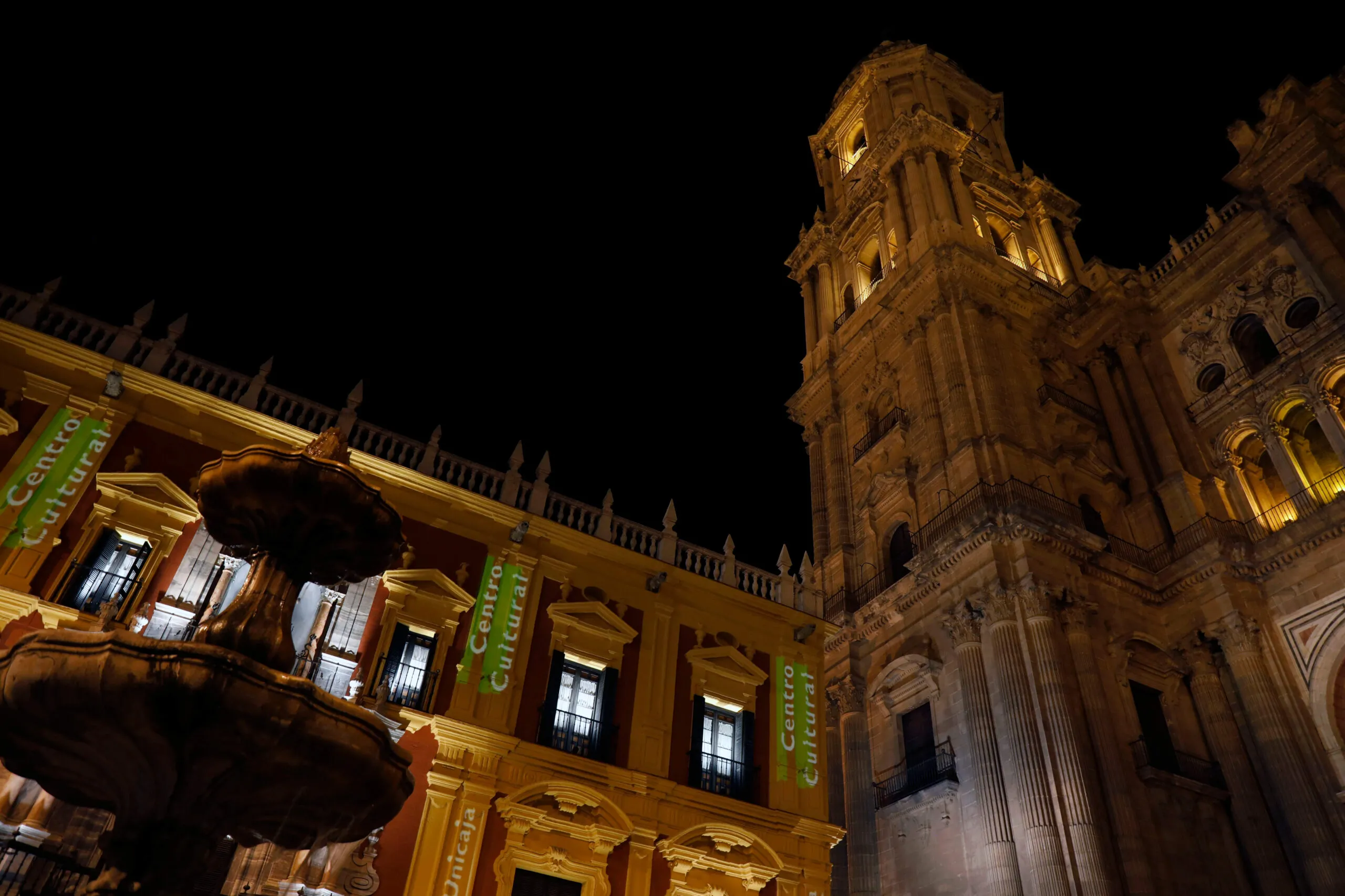 Vista nocturna del edificio del Centro Cultural con estandartes verdes colgando de su fachada barroca, junto a la torre iluminada de la Catedral de Málaga bajo un cielo oscuro.
