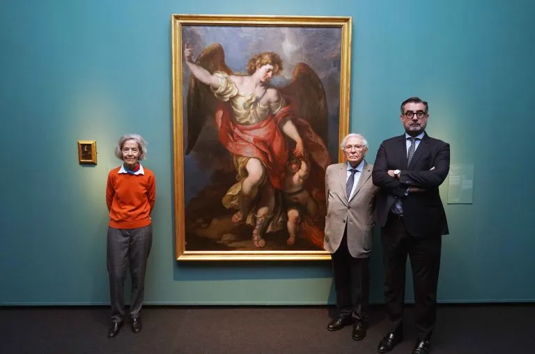 Tres personas de pie frente a un gran cuadro enmarcado de una figura angelical con alas vestida con túnicas rojas, expuesto en una pared de color verde azulado en una galería