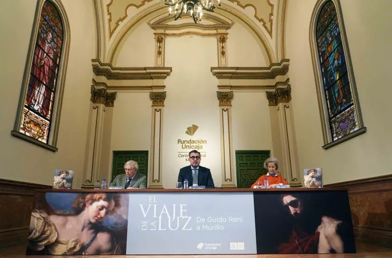 Tres personas sentadas a una mesa larga durante una rueda de prensa dentro de un salón ornamentado con altos ventanales de cristal tintado, un techo abovedado y una lámpara de araña, con una pancarta que reza 'El Viaje de la Luz' y el logotipo de la Fundación Unicaja visibles en la pared detrás de ellos.