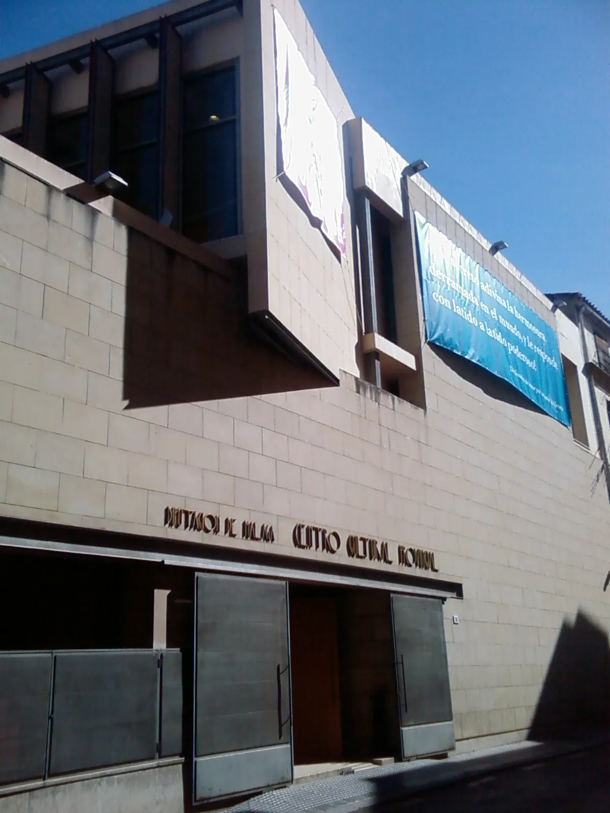 Fachada exterior del edificio del Centro Cultural Provincial con revestimiento de piedra beige, arquitectura angular, puertas de entrada y una pancarta azul colgada en la pared superior bajo un cielo azul despejado.