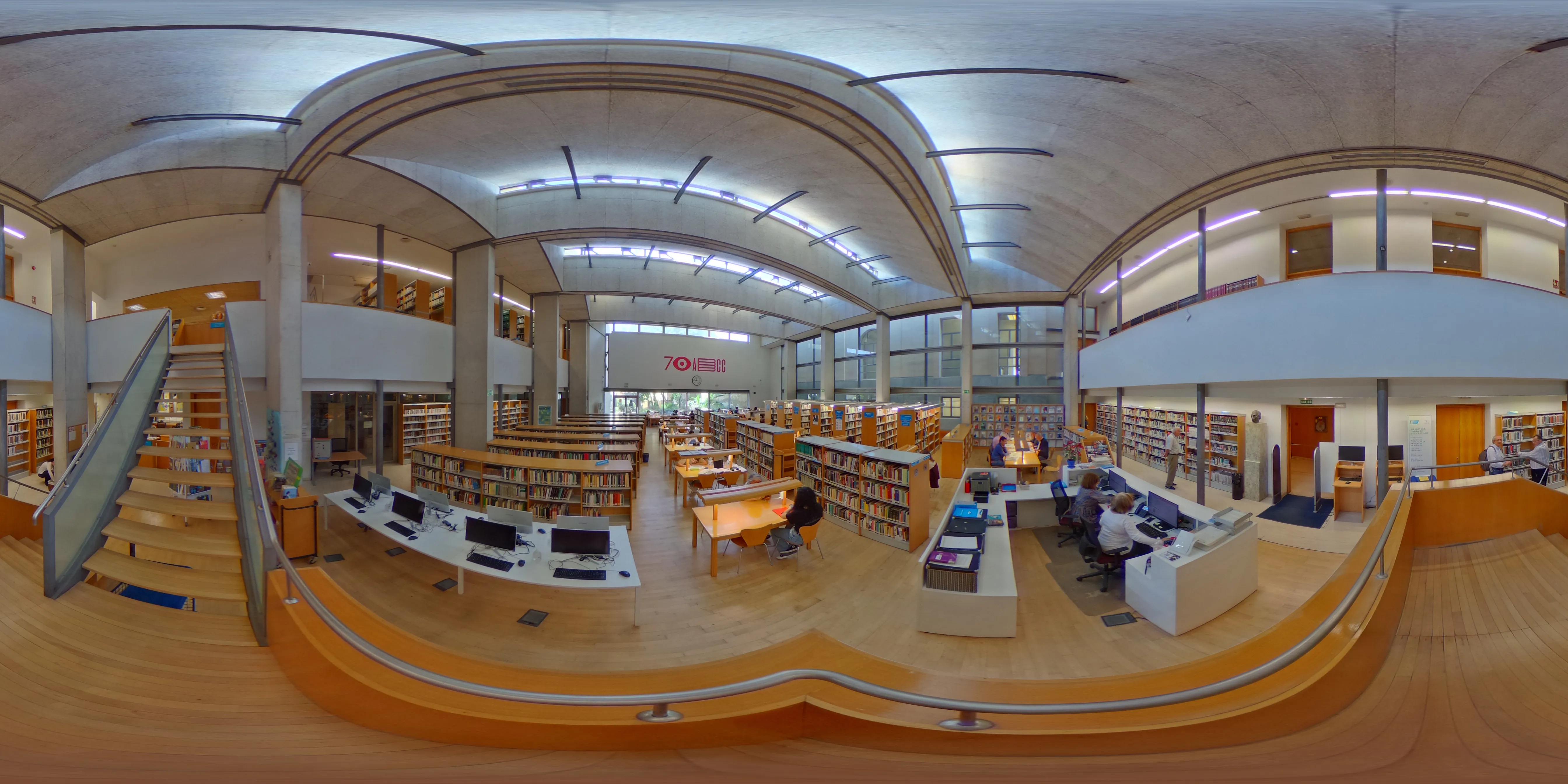 Vista interior panorámica de una biblioteca con techo curvo, estanterías, mesas de lectura, estaciones de ordenador y usuarios, dentro del Centro Cultural María Victoria Atencia