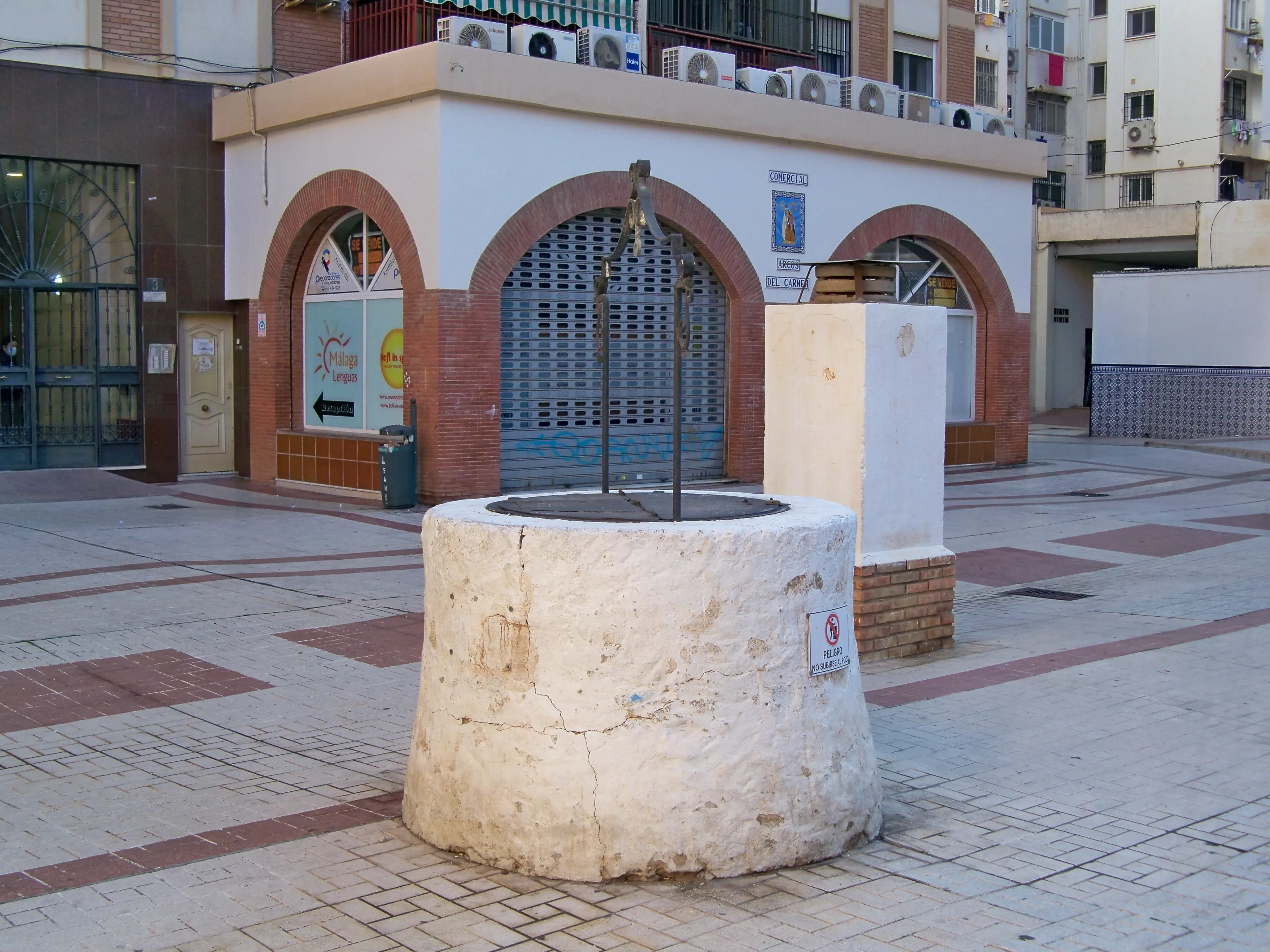 Pozo circular de piedra con marco de metal situado en una plaza pavimentada, con la fachada de un edificio de arcos de ladrillo y bloques de apartamentos al fondo.