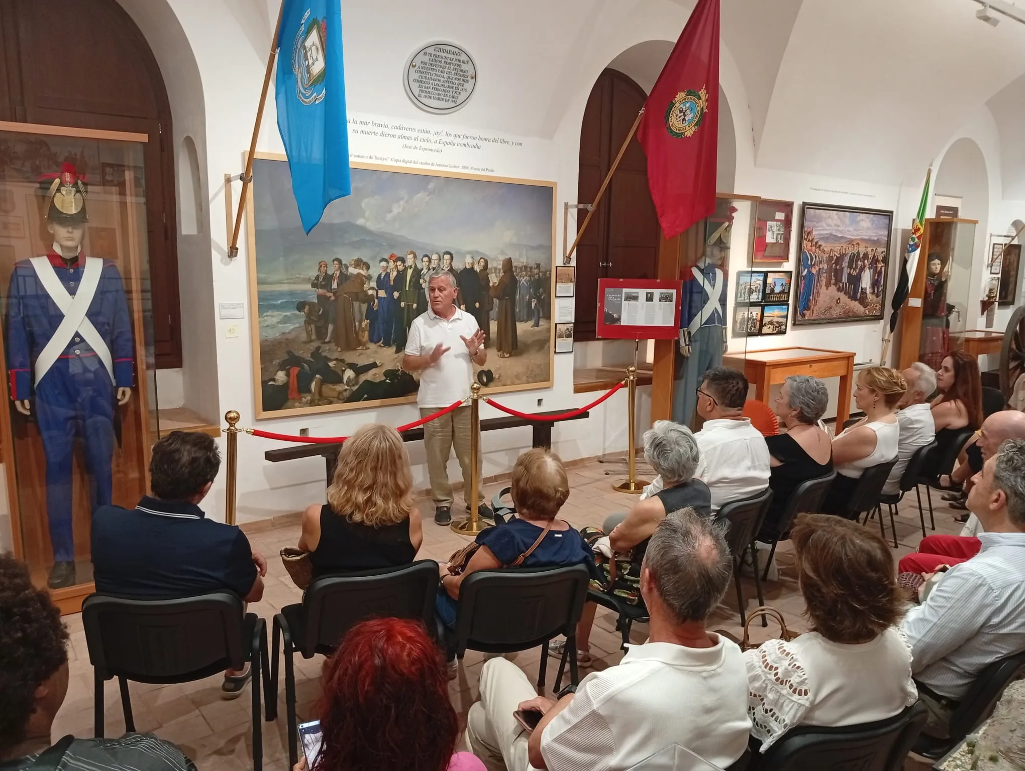 Visitantes sentados en sillas escuchando a un guía en una sala de museo con puertas arqueadas, pinturas históricas, banderas azules y rojas y maniquíes vestidos con uniformes militares.