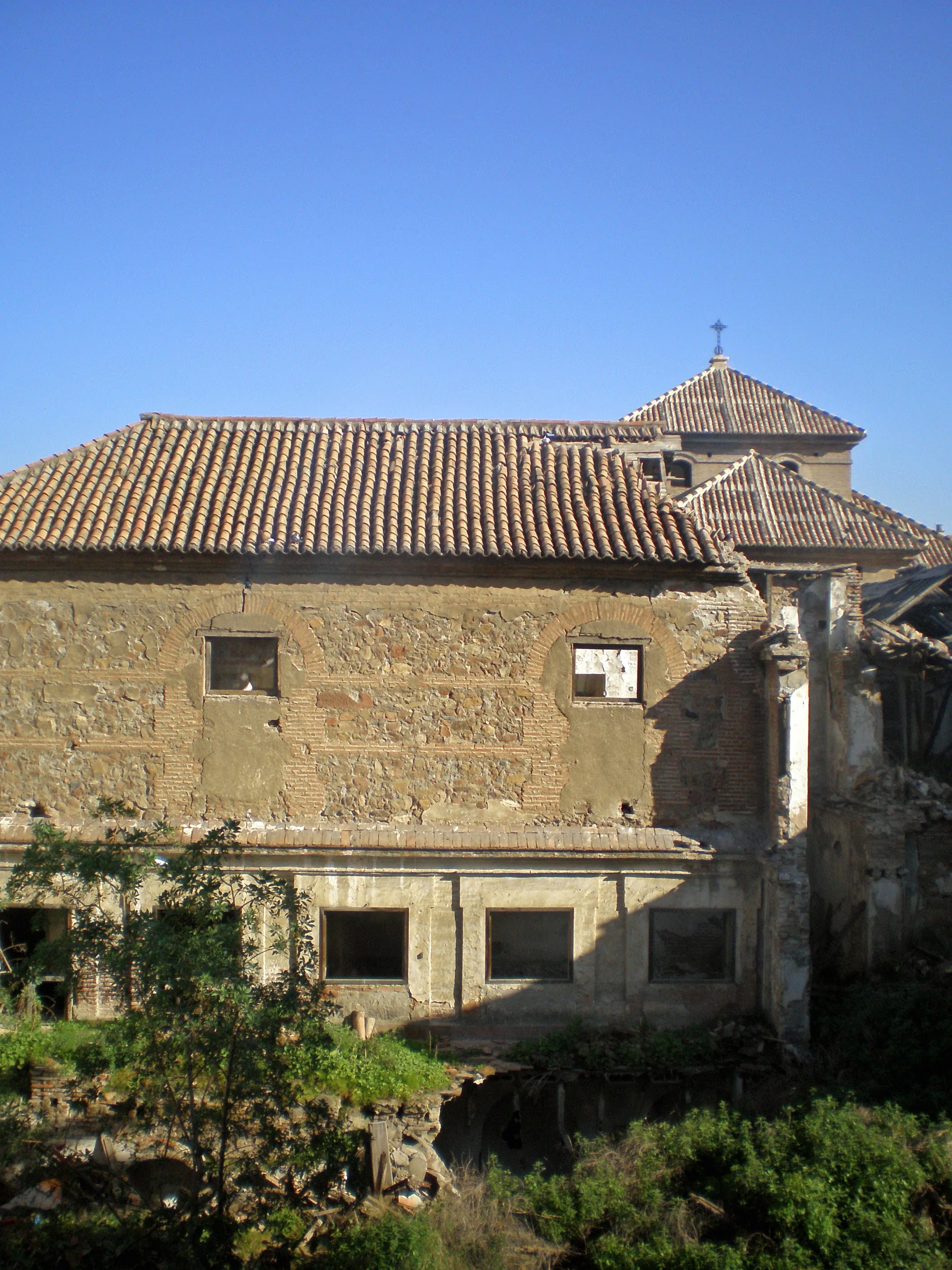 Exterior de piedra erosionada del Convento de San Andrés con tejados de terracota, una torre coronada por una cruz, ventanas rectangulares y vegetación abundante en la base bajo un cielo azul despejado.