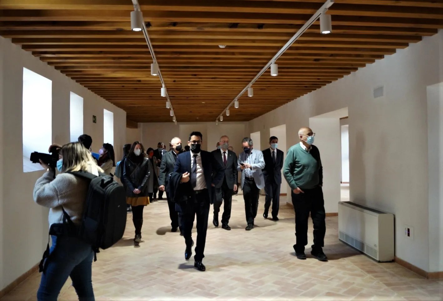 Largo vestíbulo con techo de vigas de madera, paredes blancas e iluminación en riel, con un grupo de personas caminando por el espacio.