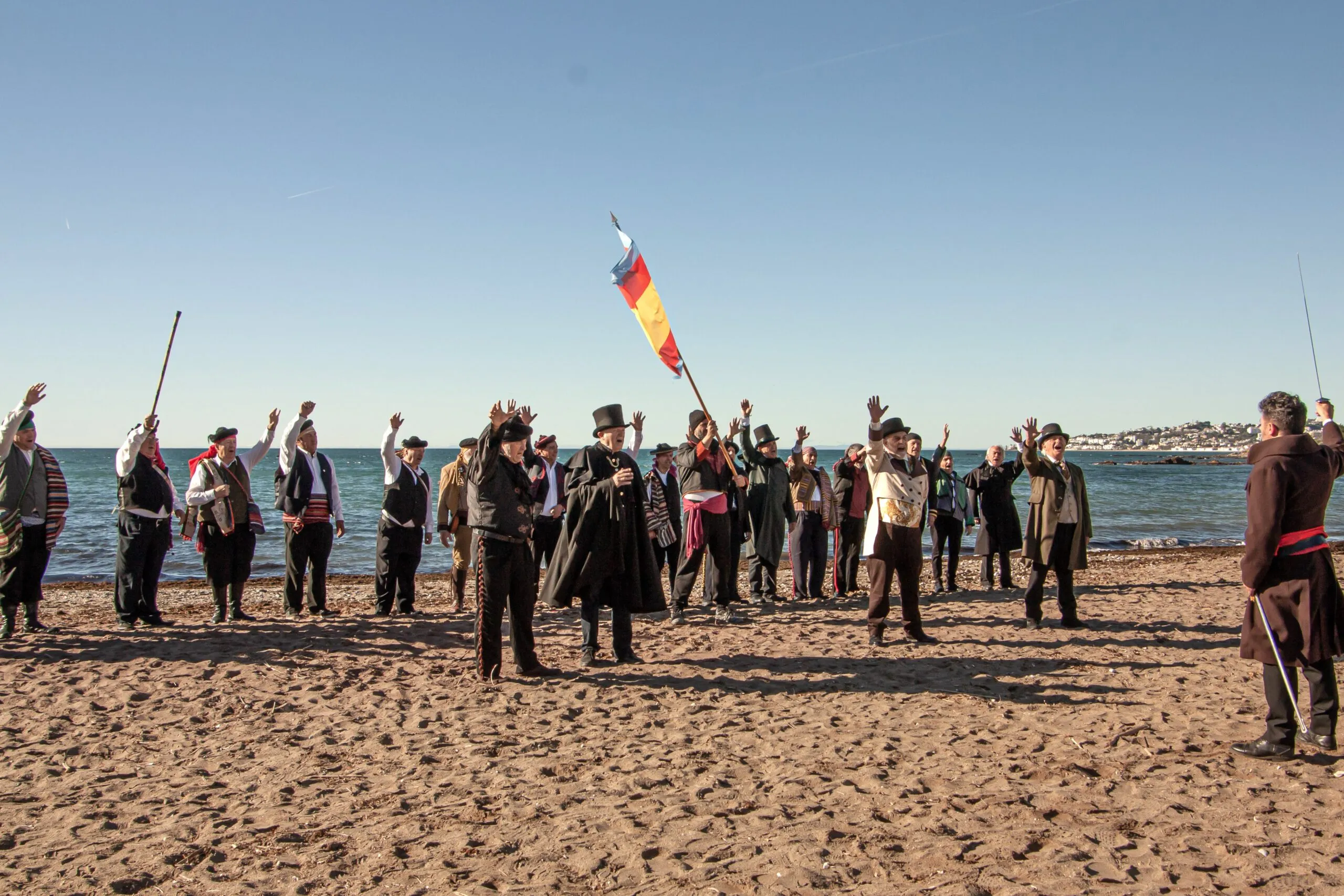 Grupo de personas con uniformes militares del siglo XIX en una playa de arena, con una persona sosteniendo una bandera española durante una recreación histórica.