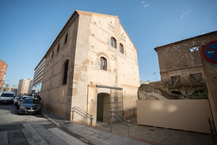 Vista exterior de un edificio histórico de piedra clara con ventanas arqueadas y una entrada con rampa y barandillas de metal, coches aparcados a lo largo de la calle y una ruina parcial visible a la derecha.