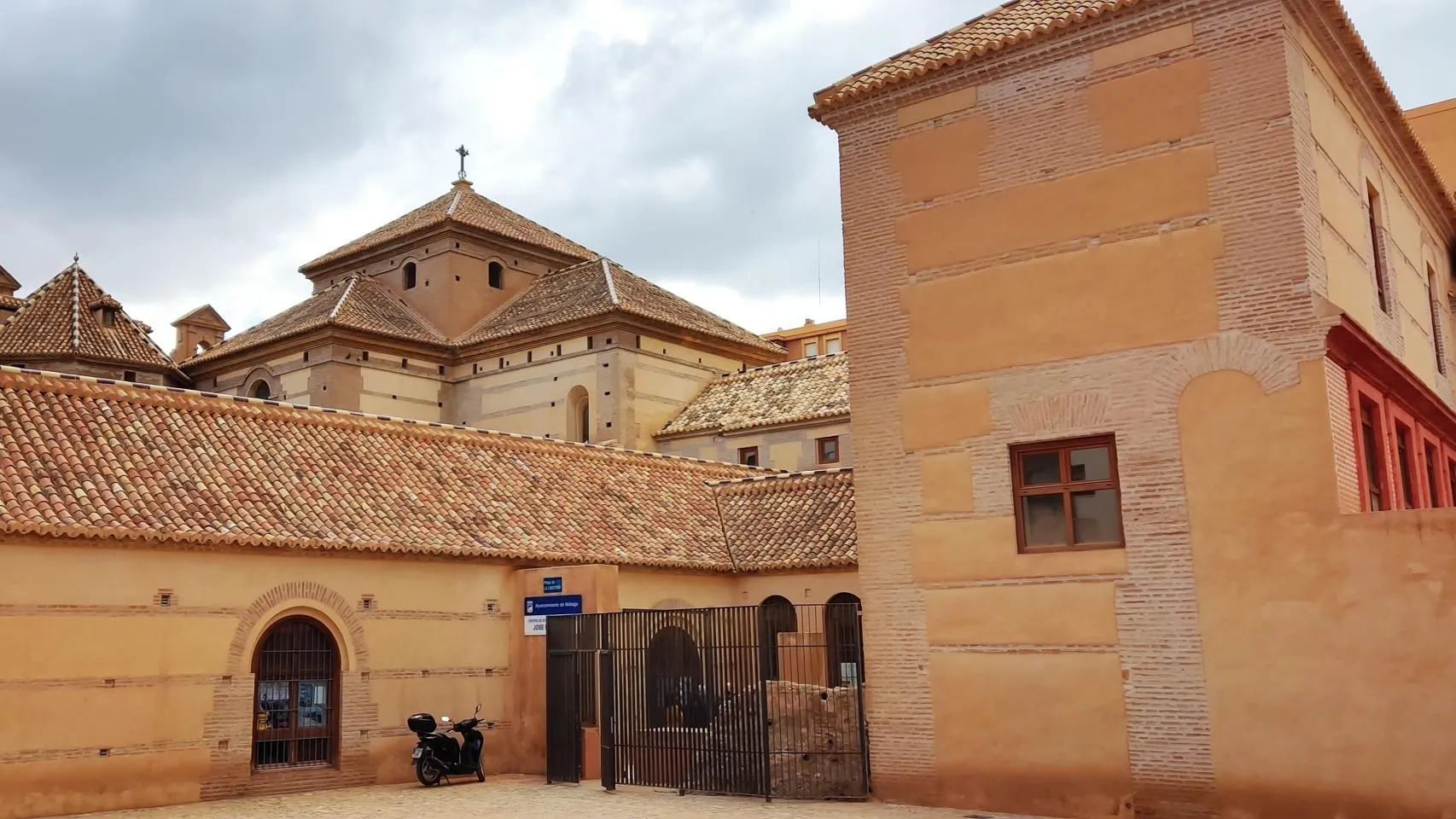 Vista exterior de un complejo de edificios históricos con paredes de terracota, tejados de tejas, una entrada arqueada y una puerta de metal negro.