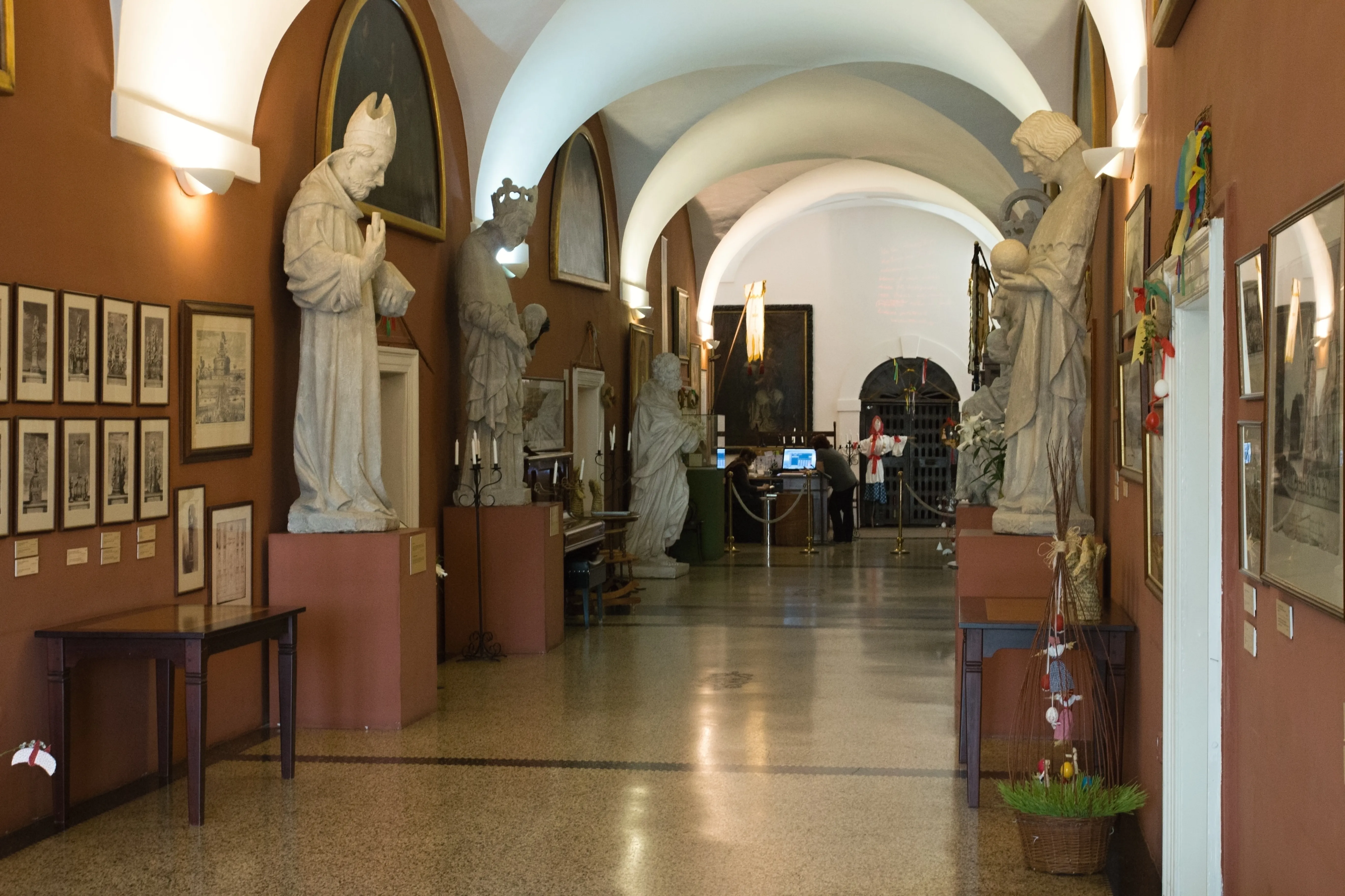 Interieur hal van een museum met gewelfde plafonds, witte beelden en ingelijste foto's aan bruine muren